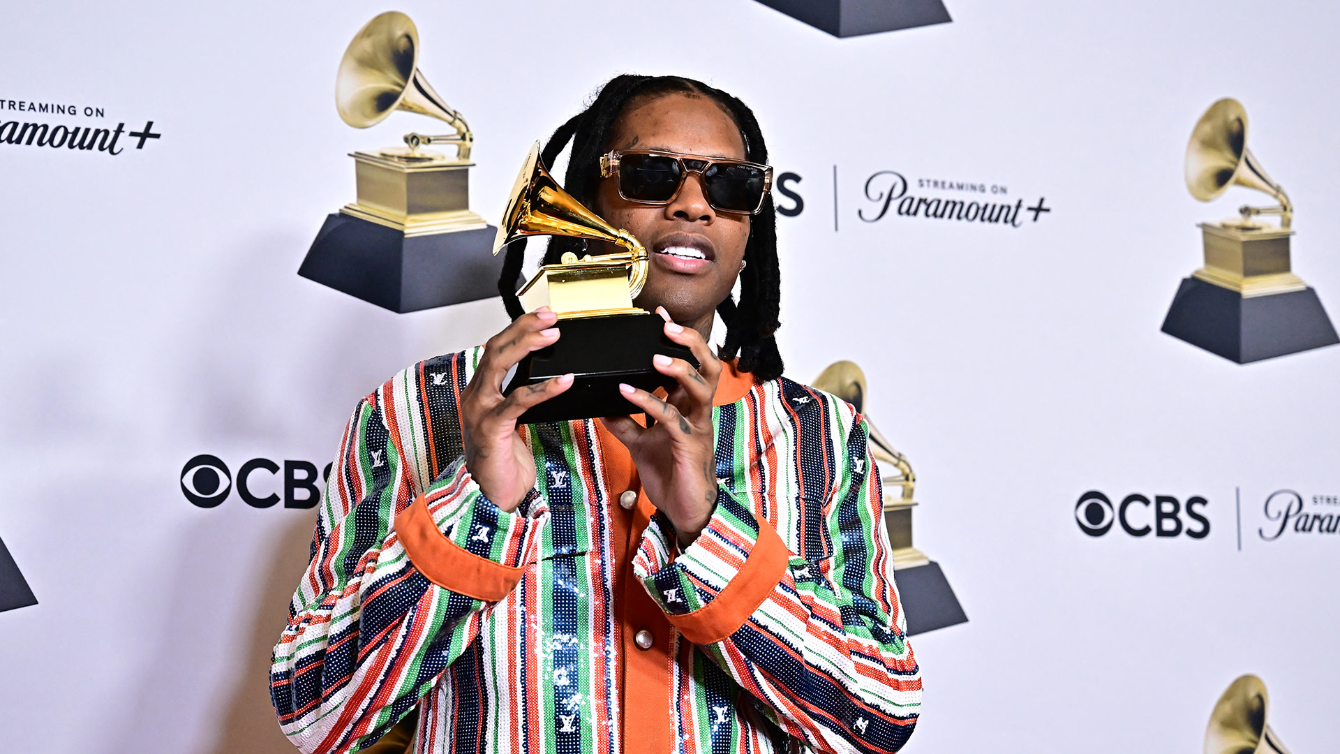 Lil Durk in a colorful striped jacket holds a Grammy award, posing on the red carpet with logos for CBS and Paramount+ in the background.