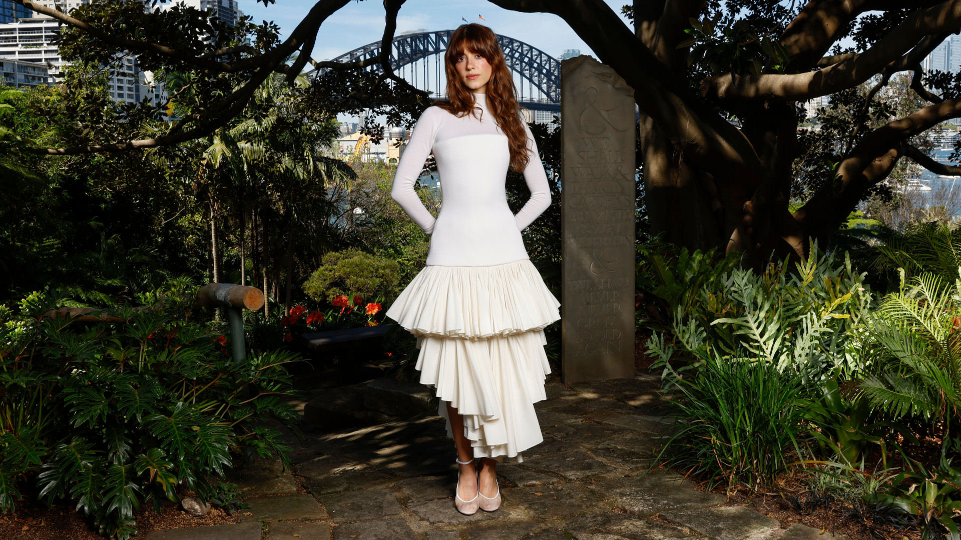 Daisy Edgar-Jones in a white outfit stands in a garden, with a view of a bridge and city skyline in the background.