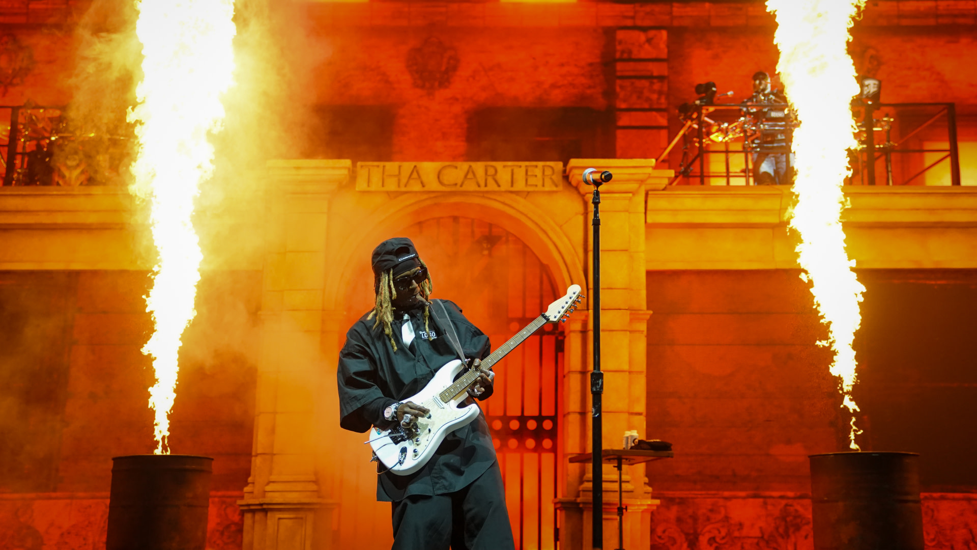 Lil Wayne performs on stage with a guitar, surrounded by flames, in front of a backdrop labeled "Tha Carter."