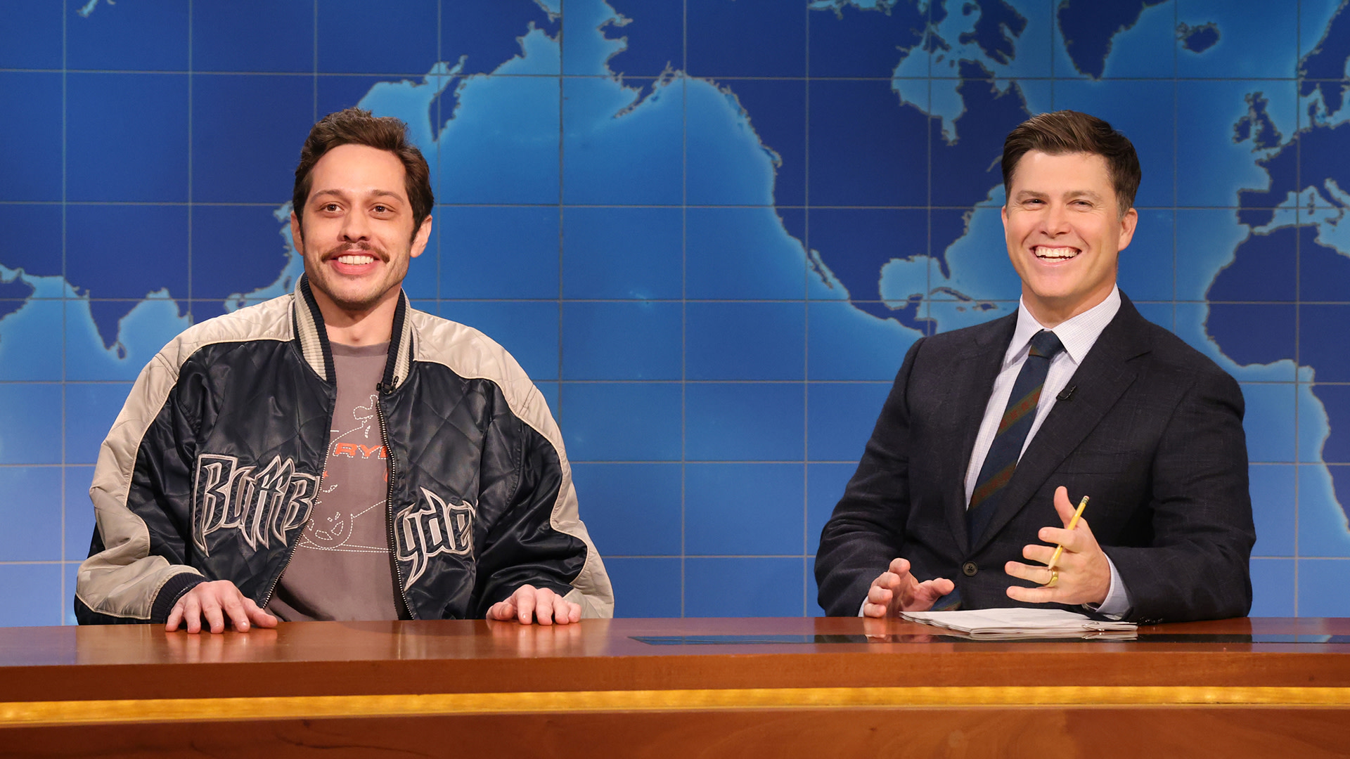 Two men sitting at the 'Saturday Night Live' Weekend Update news desk. Pete Davidson, on the left, wears a casual jacket, and Colin Jost, on the right, is in a suit. They are both smiling.