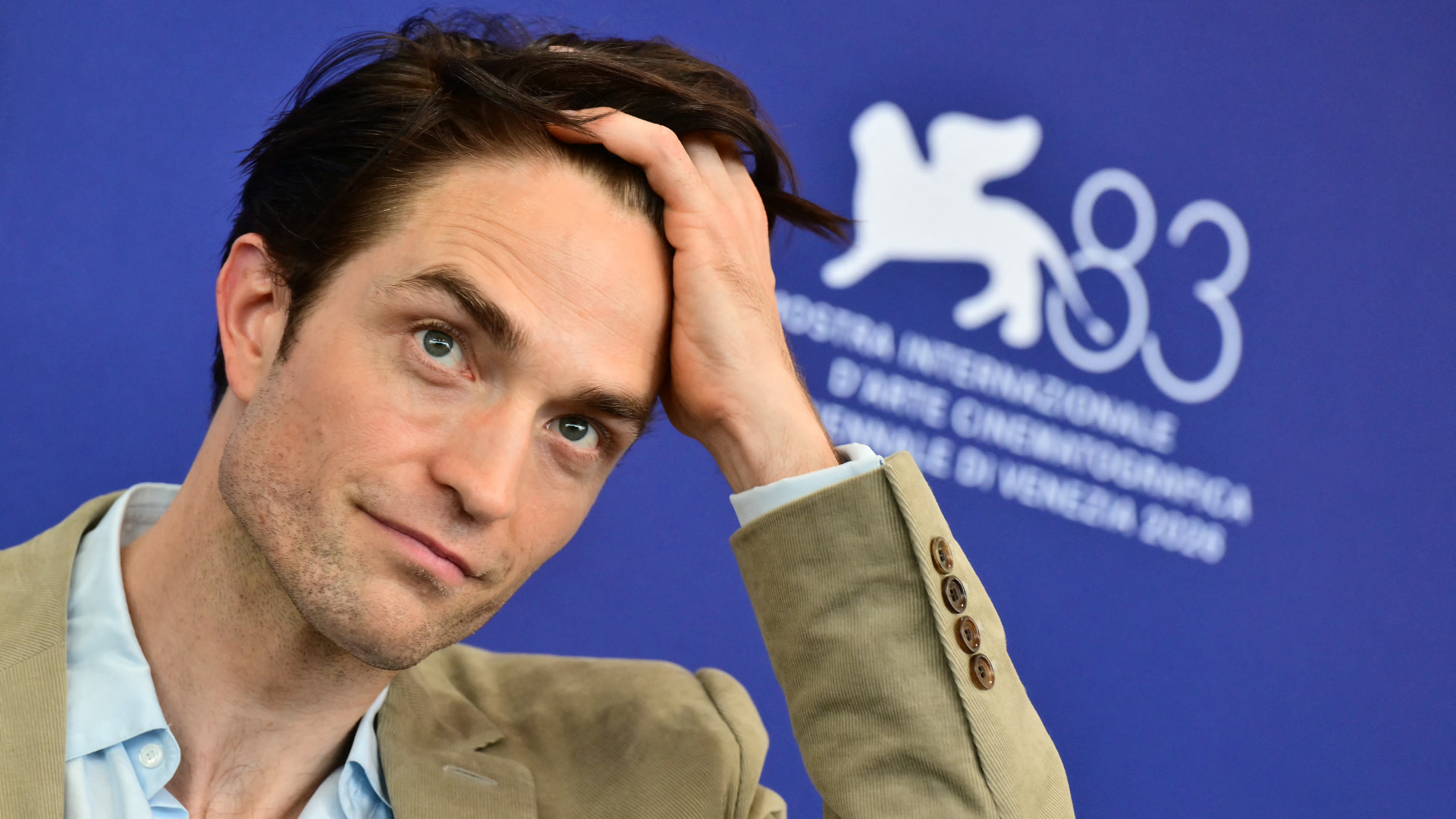 Robert Pattinson at a press event, wearing a beige suit and light blue shirt, with a blue background featuring the Venice Film Festival logo.
