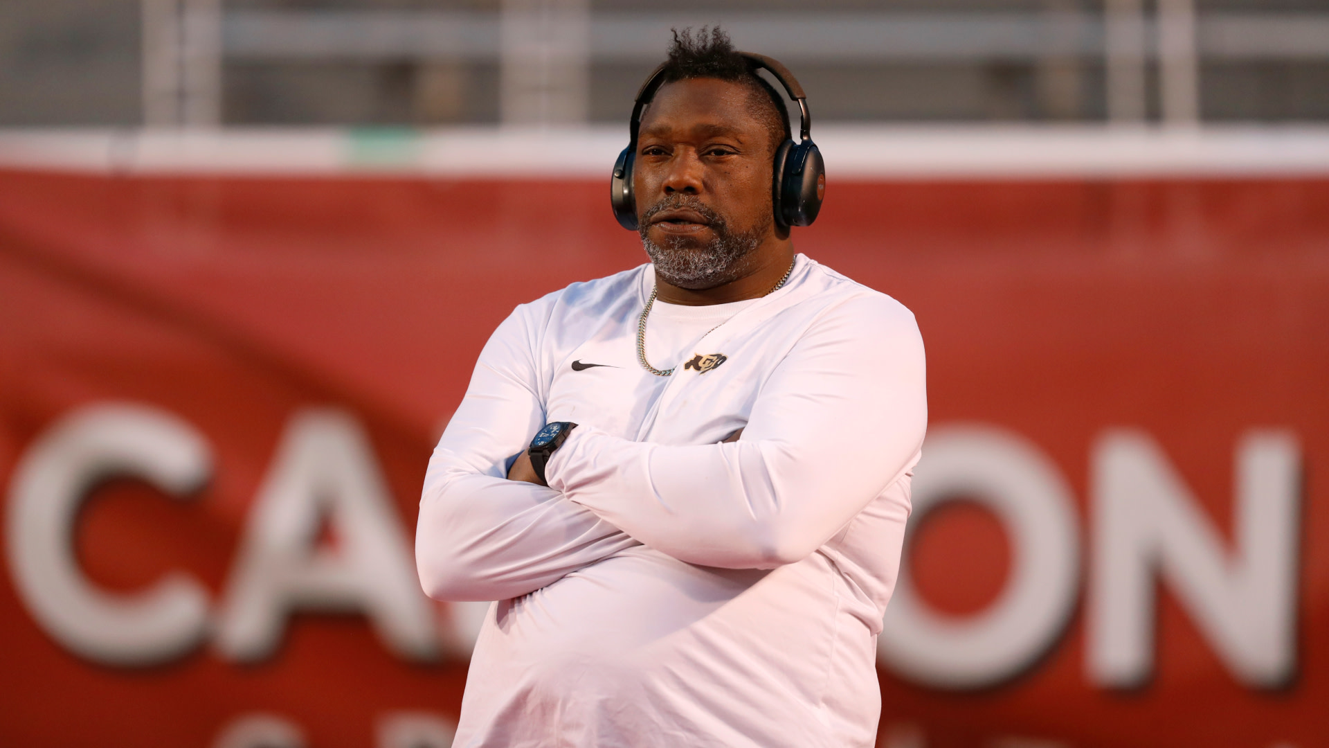 Warren Sapp in a white shirt with headphones stands with crossed arms, looking focused. The background is blurred with a red sign.