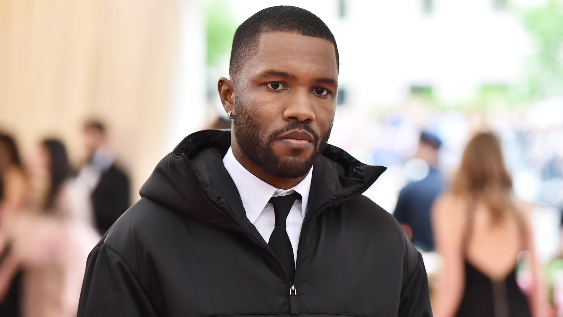 Frank Ocean wearing a black jacket and tie at a formal event, with a blurred crowd in the background.