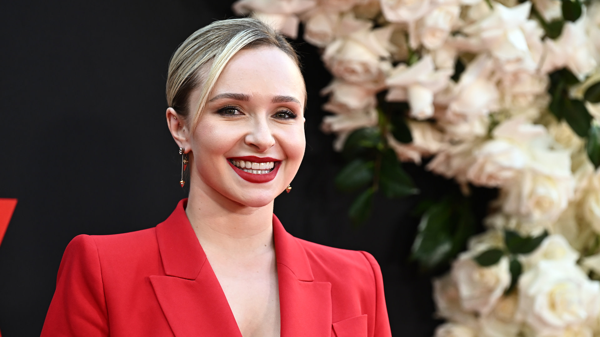 Hayden Panettiere smiles in a red blazer, with a floral backdrop.