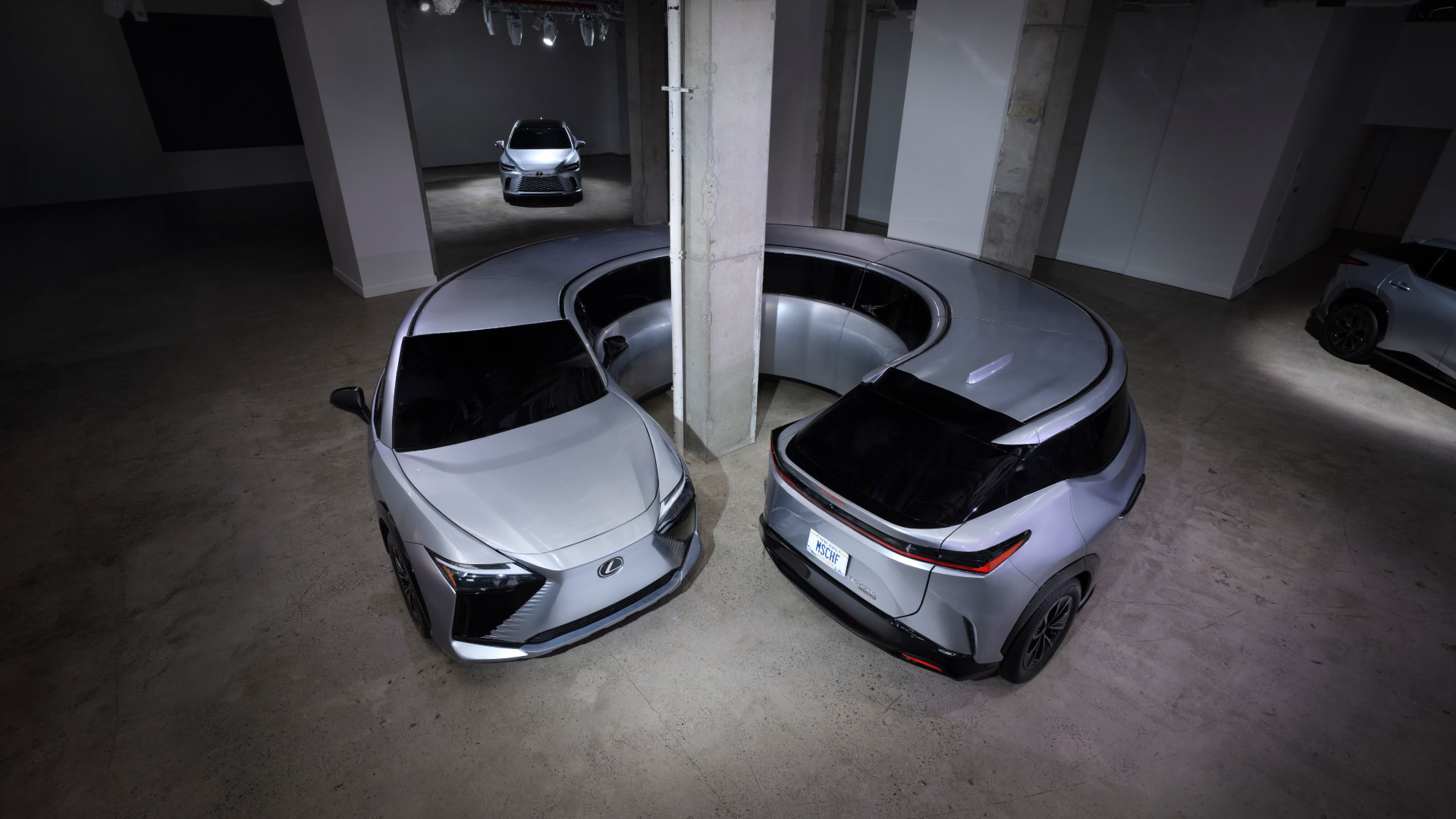 Two silver Lexus cars are parked in a showroom with a curved wall, showcasing their sleek design and modern features.