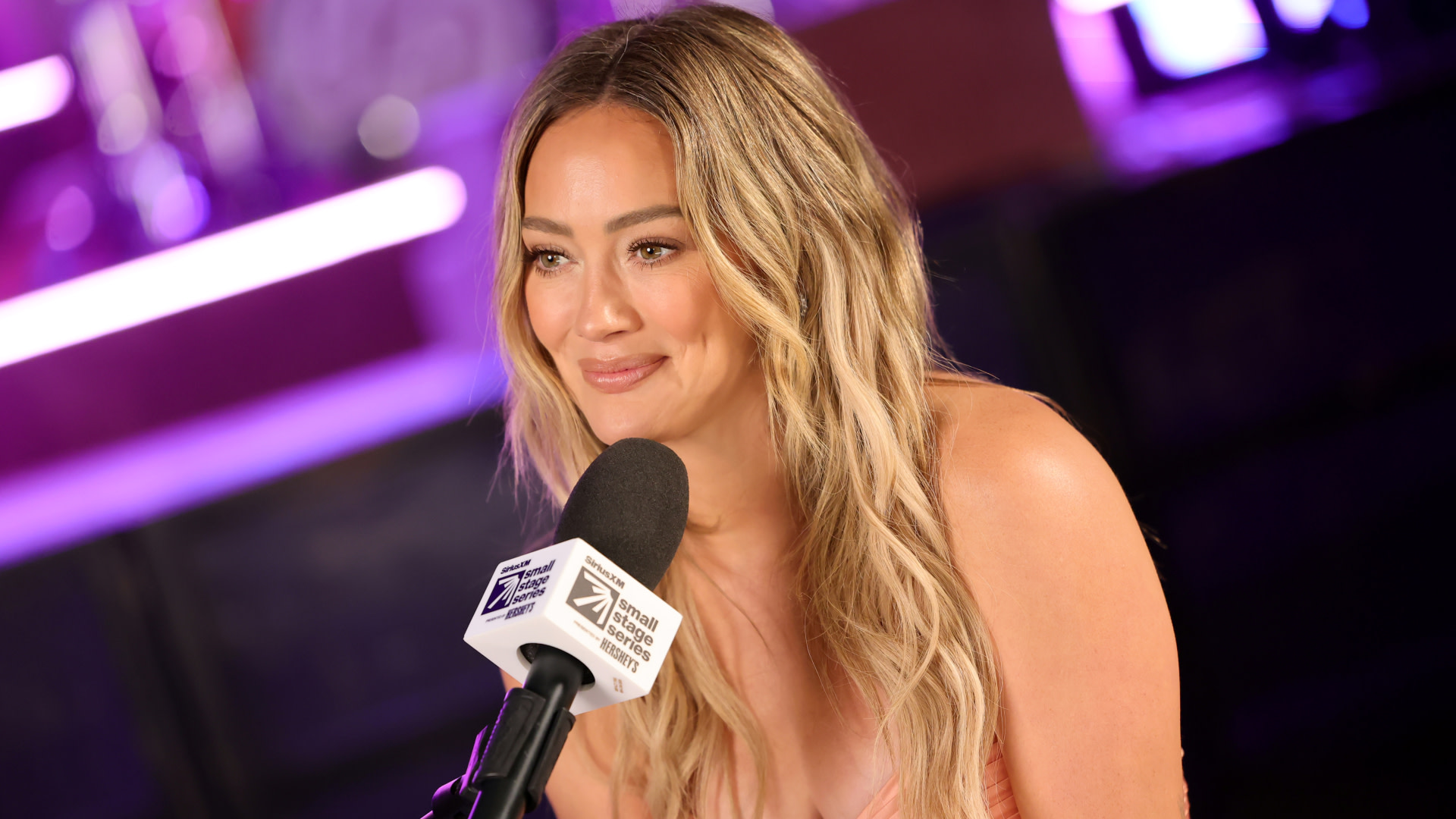 Hilary Duff with long blonde hair, smiling while speaking into a microphone at an event with purple lighting.