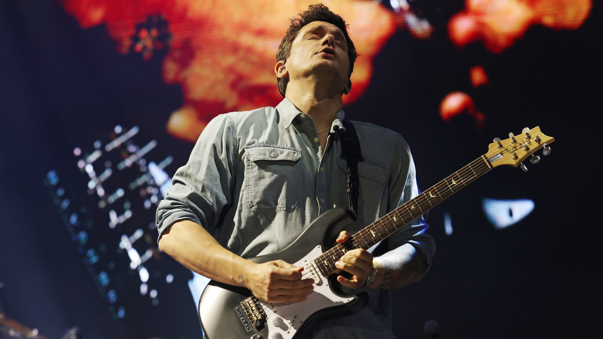 John Mayer passionately playing an electric guitar on stage, with a colorful backdrop behind him.