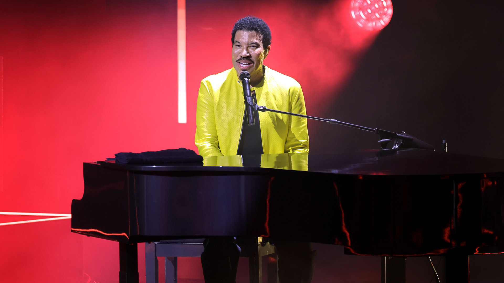Lionel Richie performing on stage, wearing a bright yellow jacket, seated at a black grand piano, with red lighting in the background.