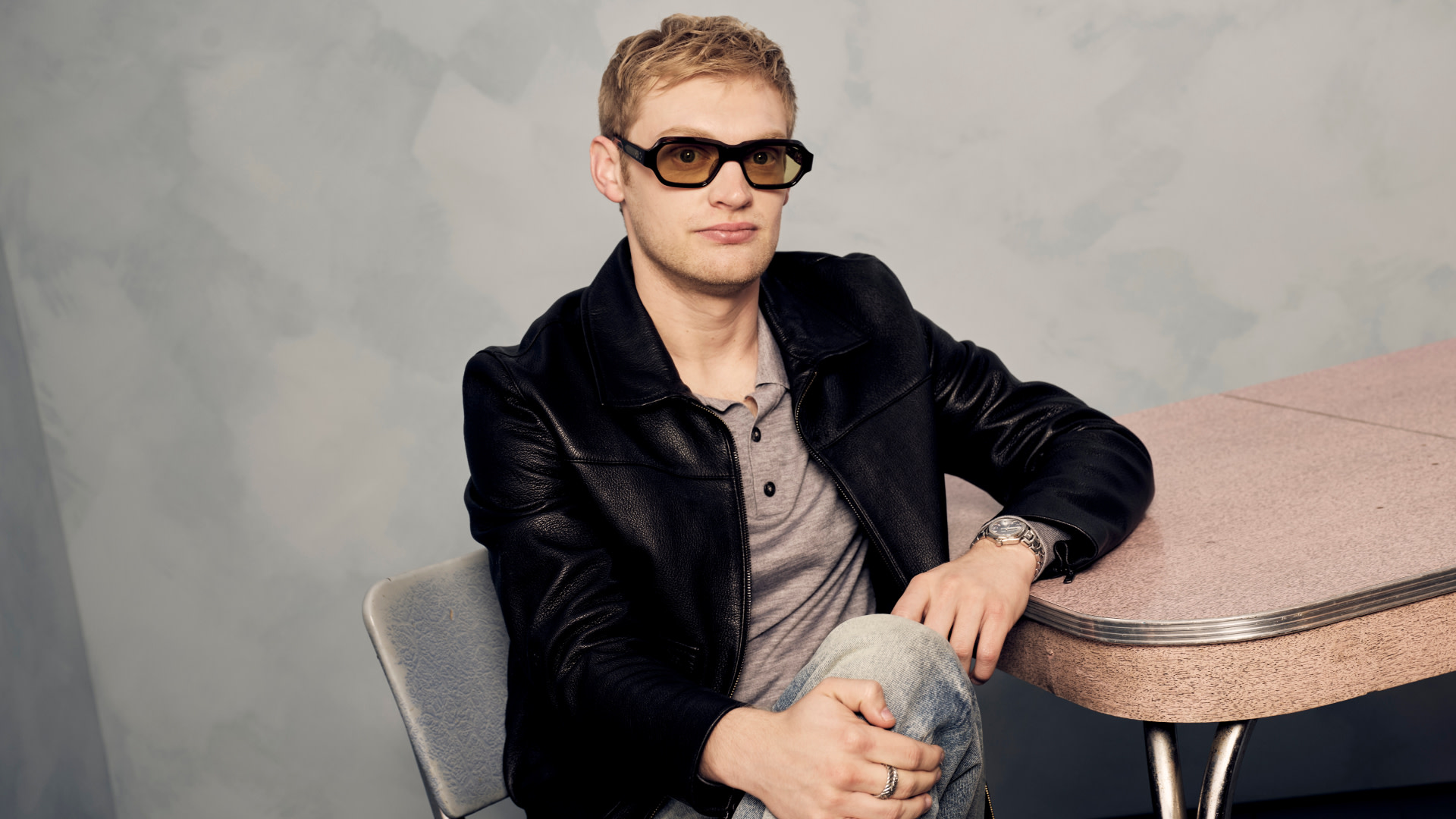 Curry Barker wearing sunglasses and a black leather jacket sits at a table, posing for a photo.