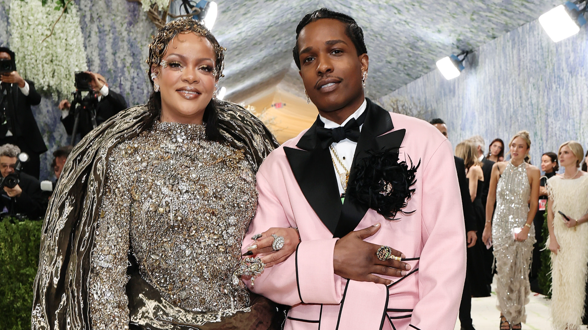 Rihanna in a shimmering gown and A$AP Rocky in a pink suit at a glamorous event, posing together with smiles.