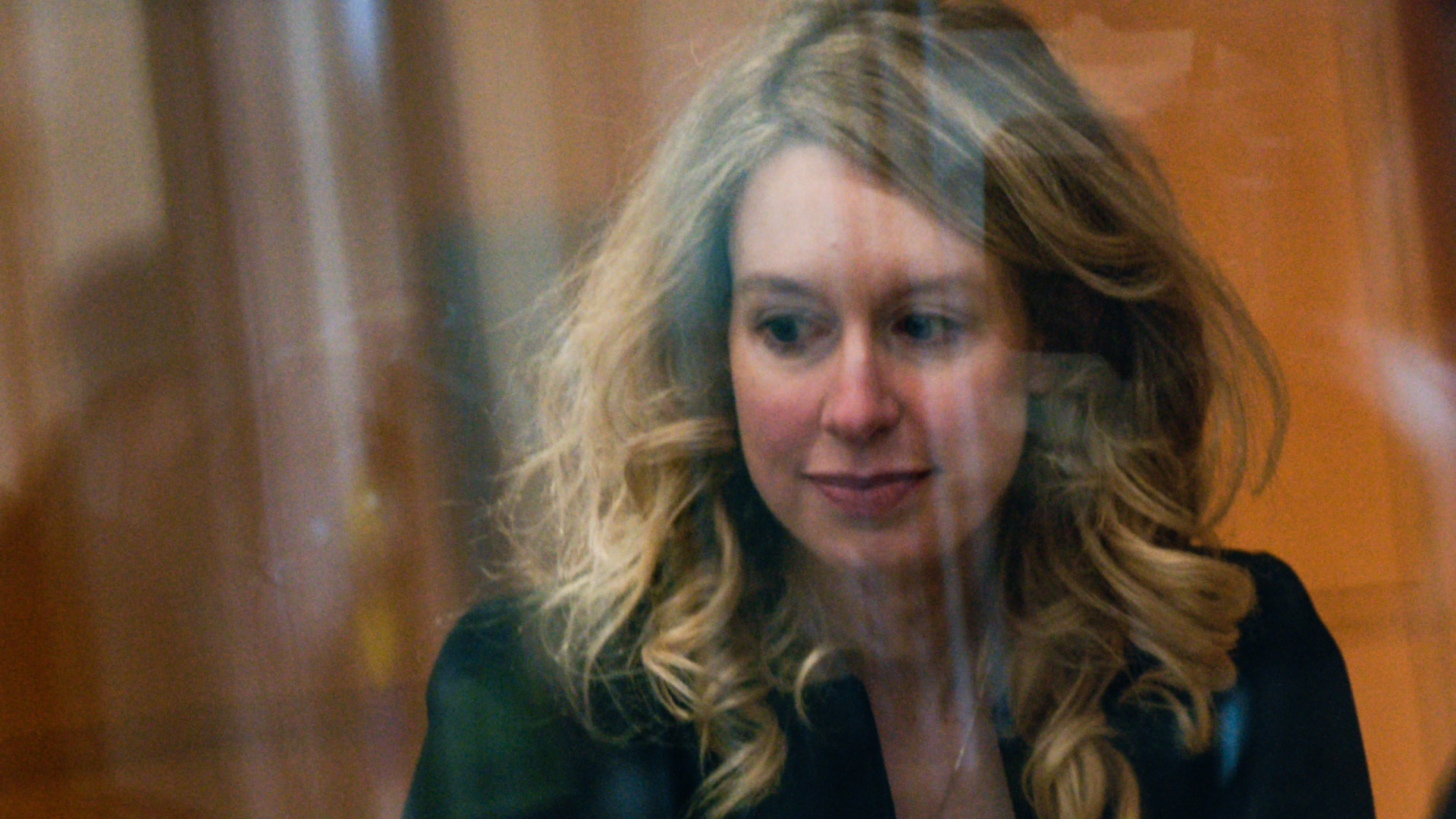 Elizabeth Holmes with long blonde hair is seen through a reflective surface, wearing a dark top, looking to the side.