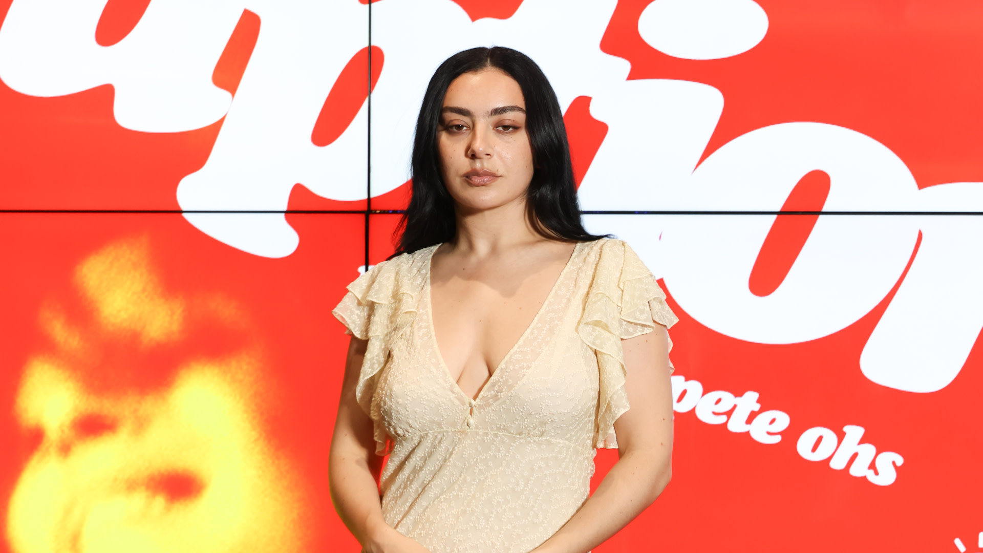 Charli xcx with long dark hair in a light dress stands in front of a red background with white text.