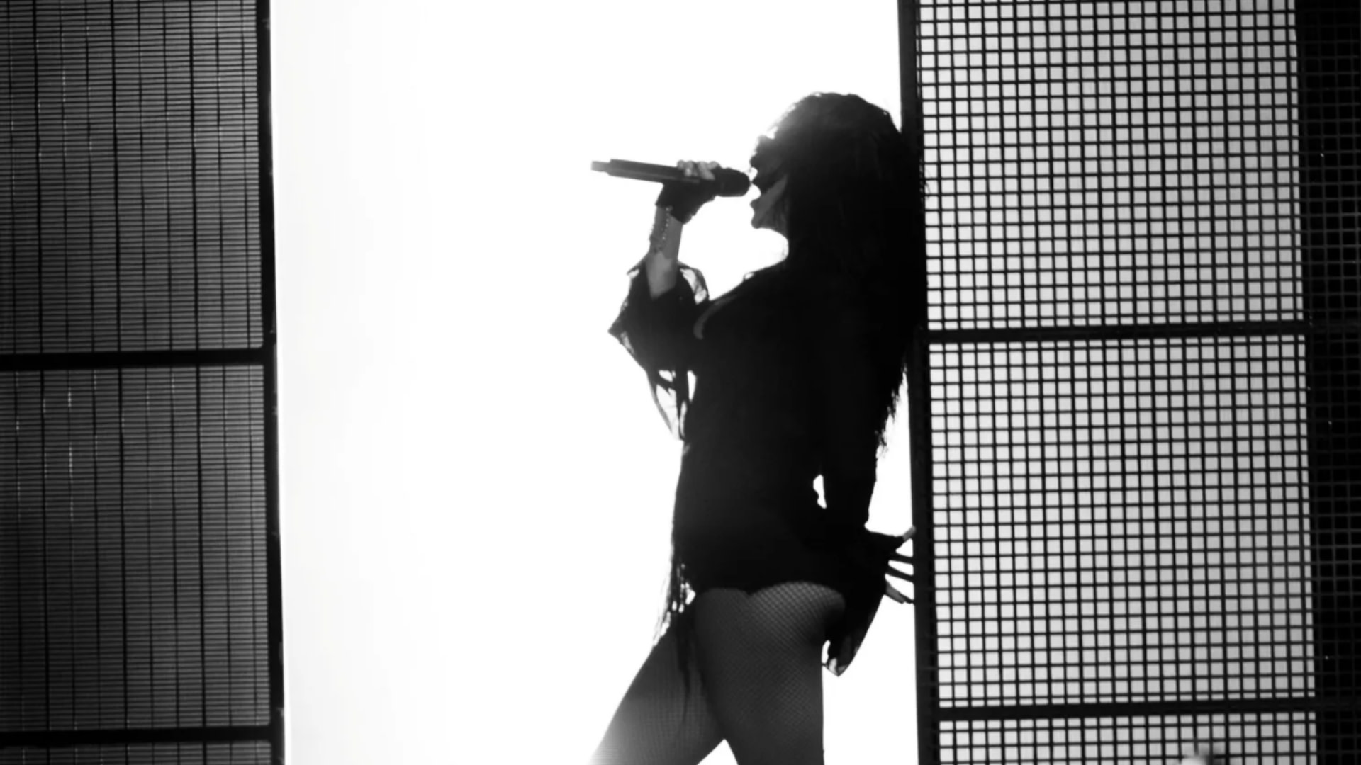 Silhouette of Charli xcx singing into a microphone on stage, with dramatic lighting and a grid-like backdrop.