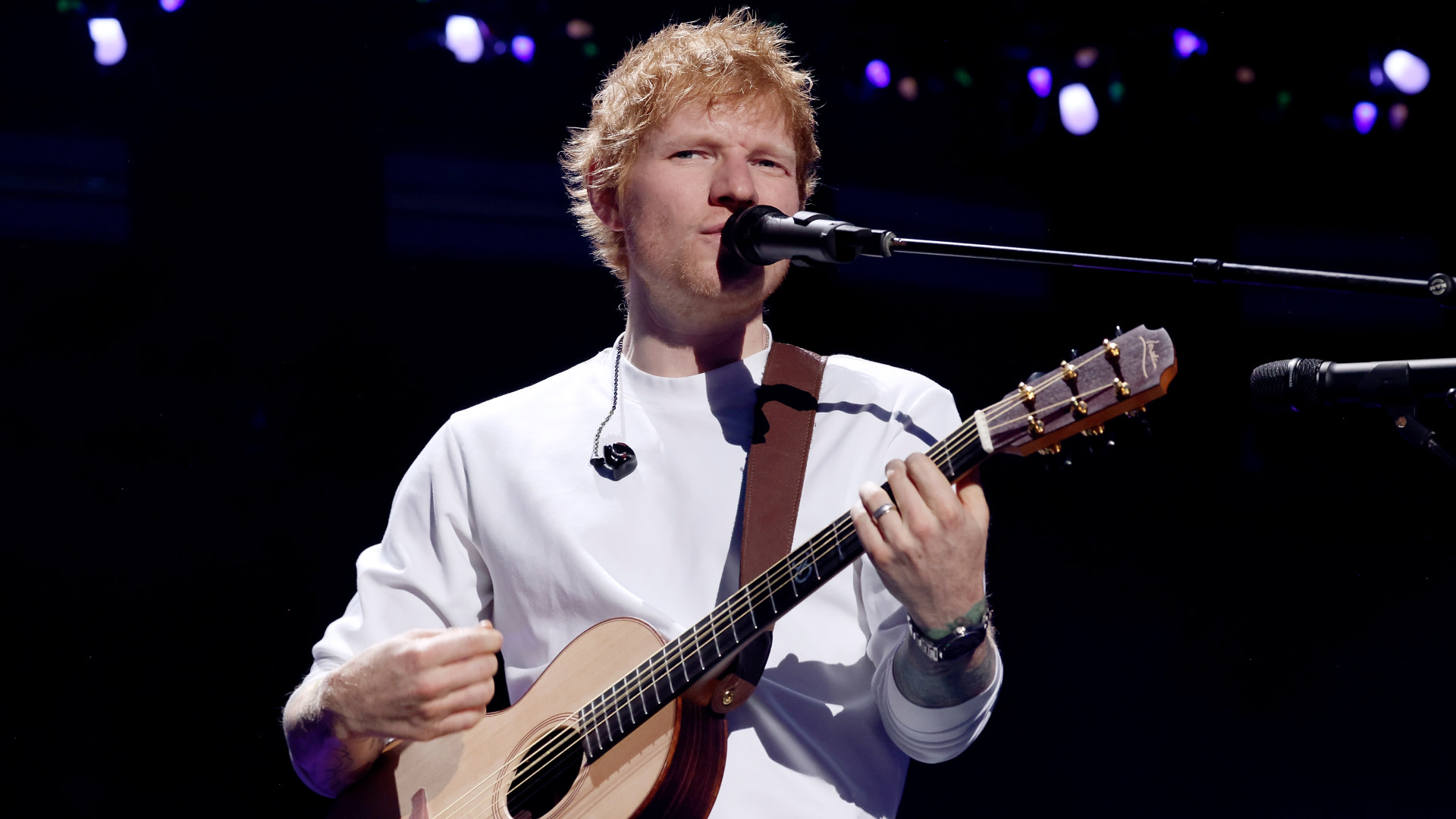 Ed Sheeran performing on stage, holding a guitar, wearing a white shirt, singing into a microphone.