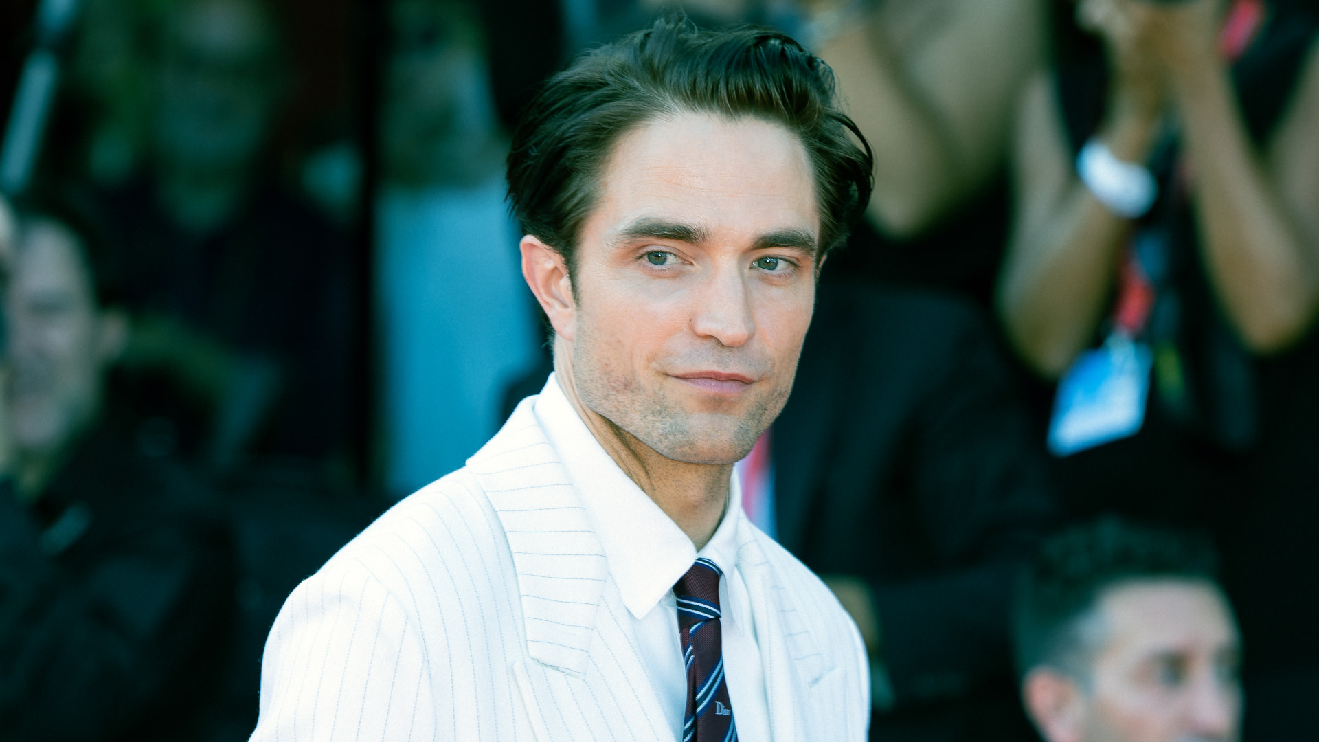 Robert Pattinson in a white suit and tie stands in a crowd, looking to the side. The background is blurred with people and cameras.