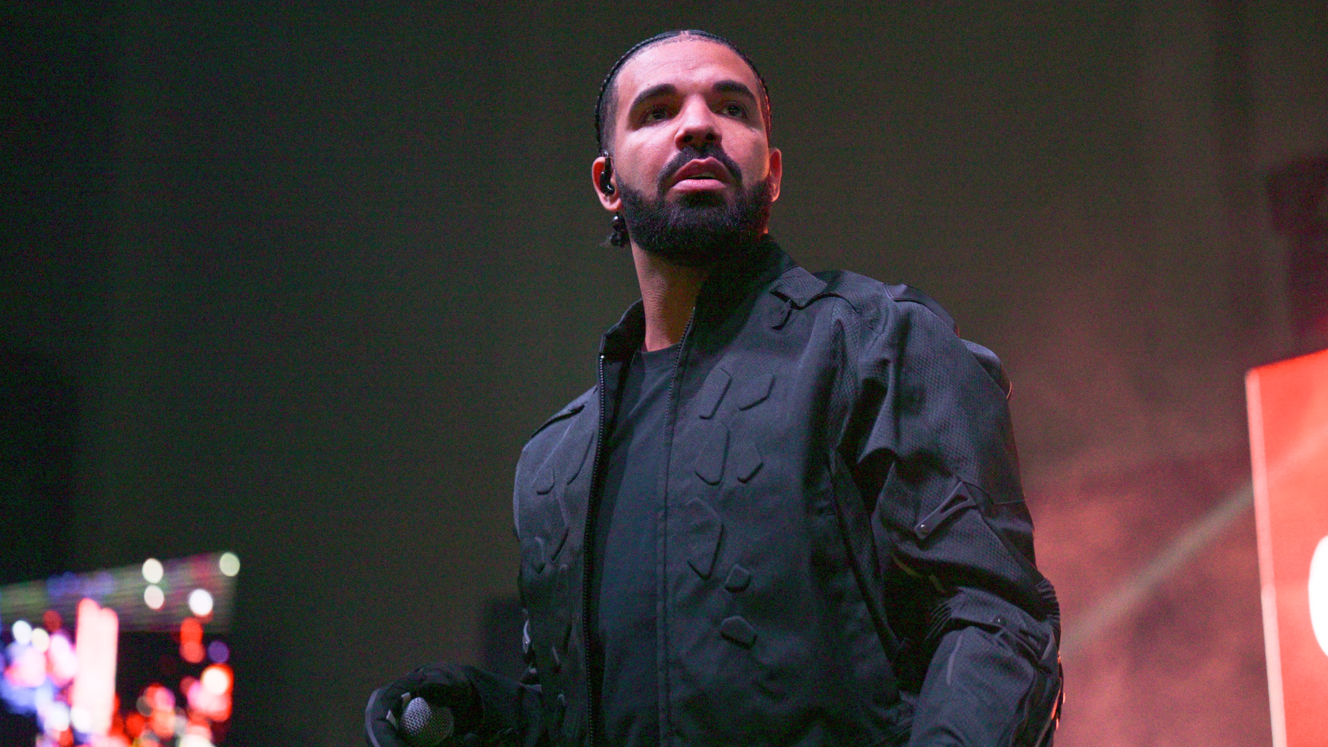 Drake performing on stage, wearing a black jacket, holding a microphone, with a focused expression.