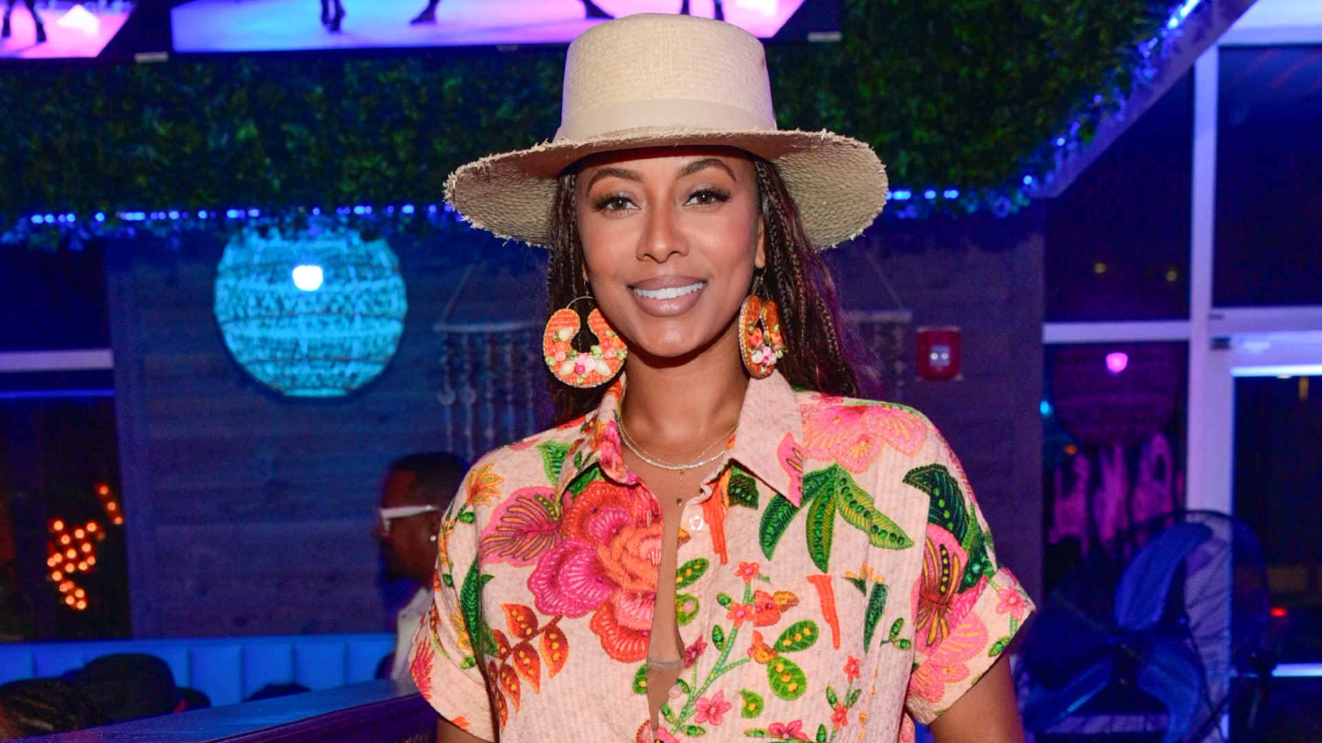 Keri Hilson wearing a floral shirt, large earrings, and a wide-brimmed hat smiles in a colorful, dimly lit setting.