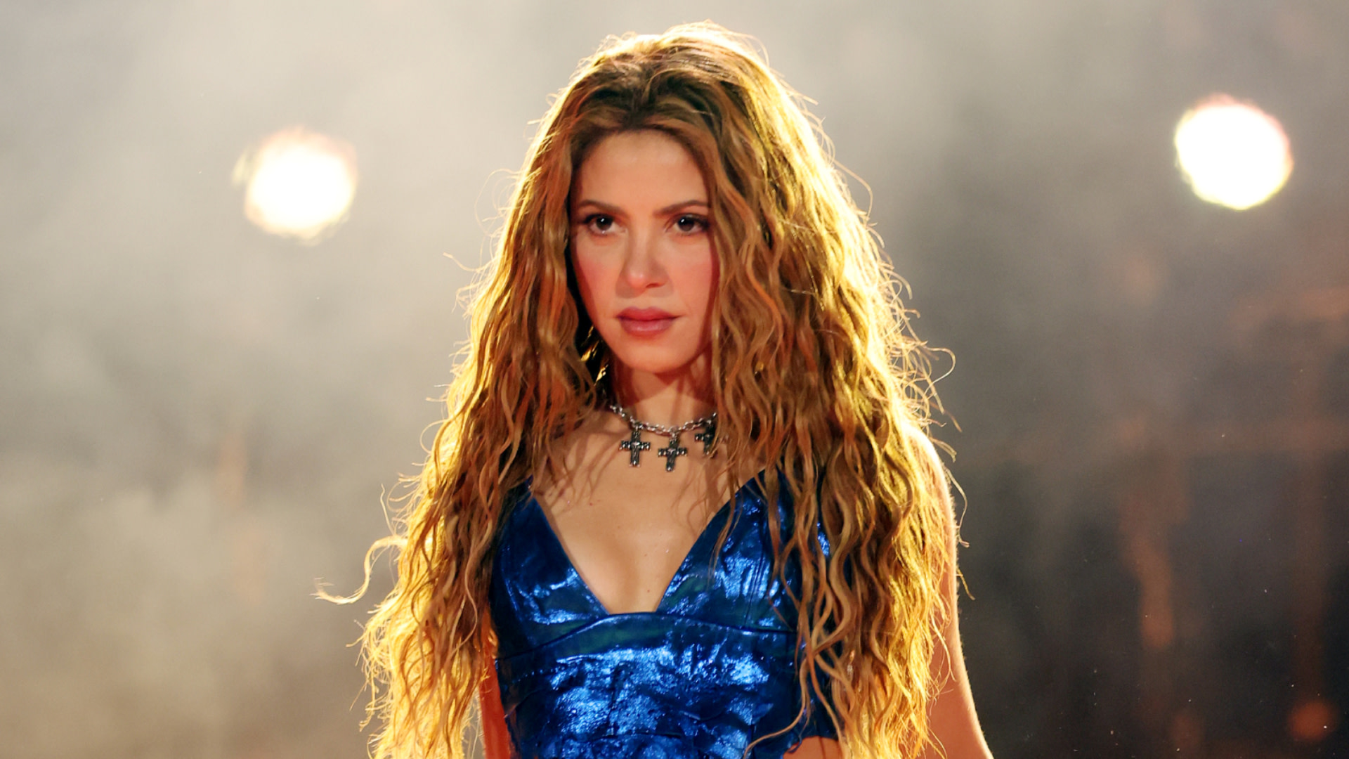 Shakira performing on stage in a blue outfit, with long wavy hair and a dramatic backdrop.