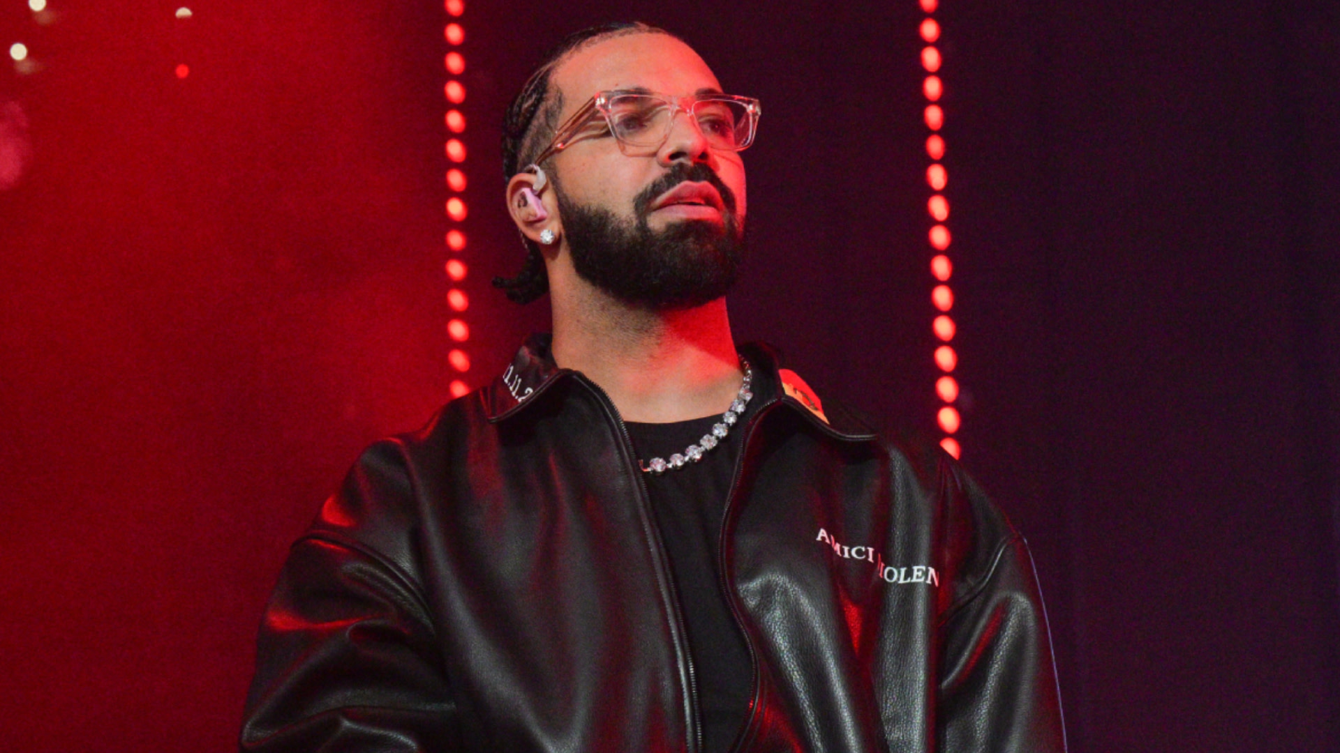 Drake with a beard and glasses wearing a black leather jacket stands on stage under red lighting.