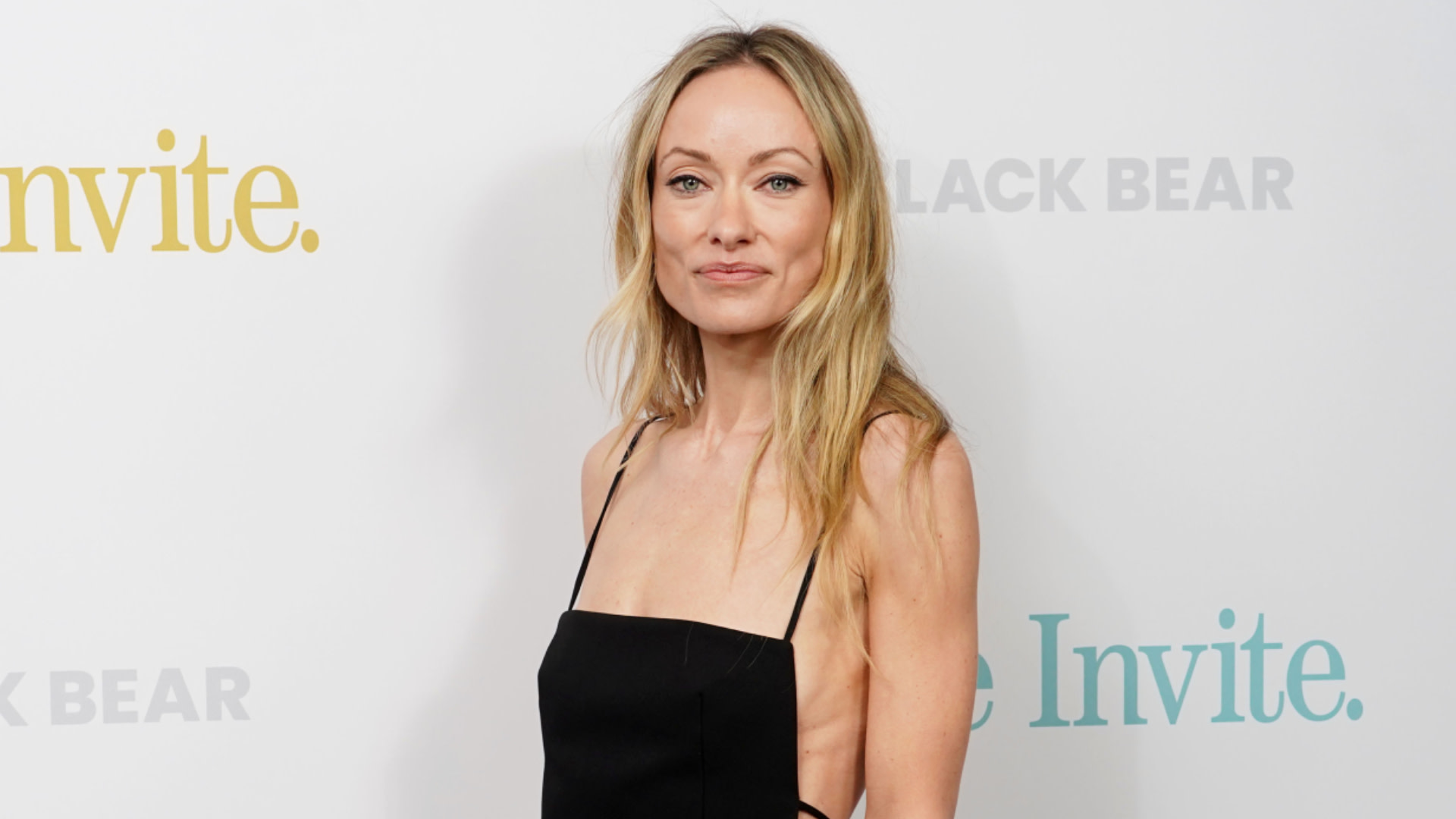 Olivia Wilde in a black dress at an event, standing against a backdrop.