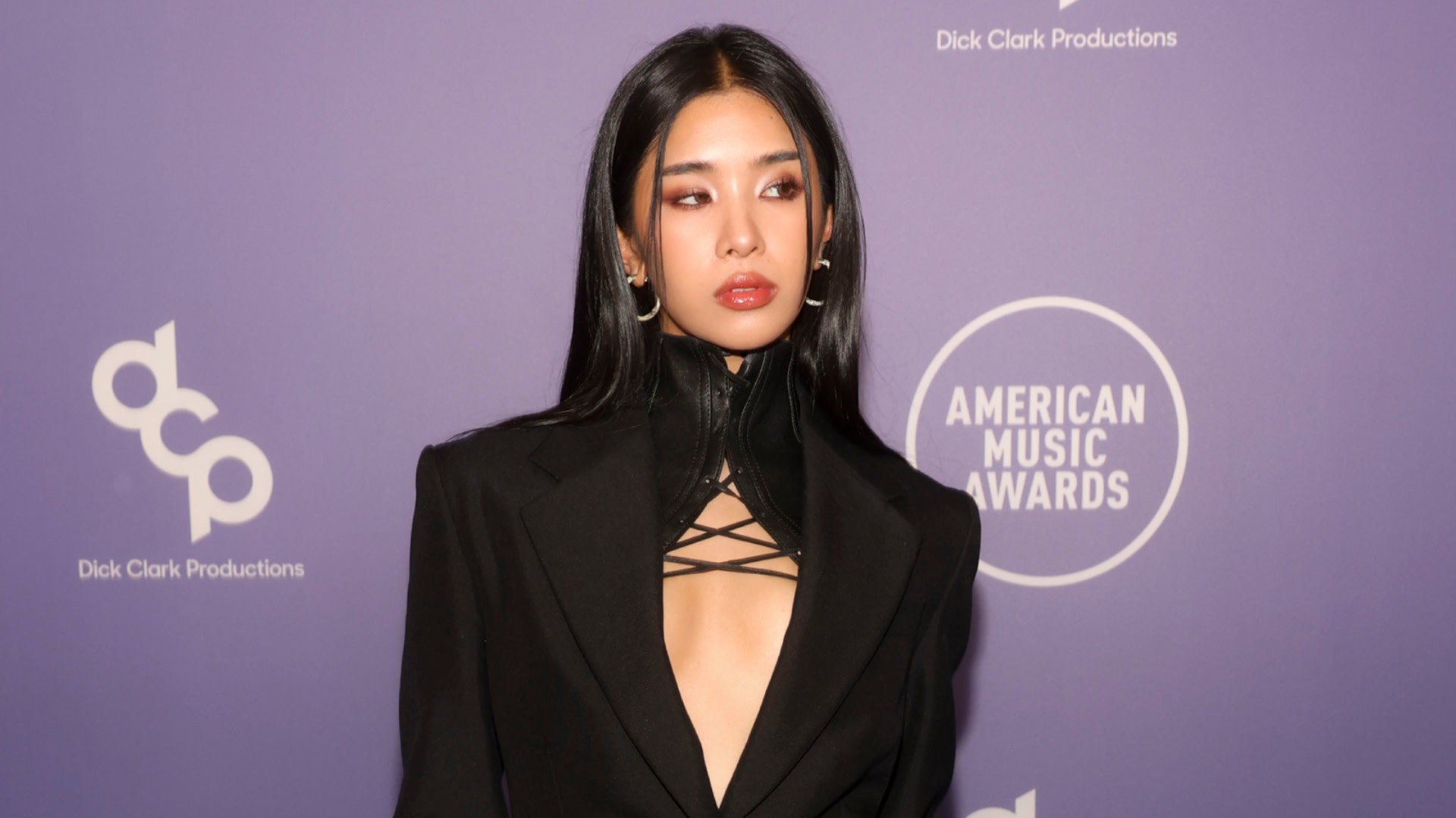 KATSEYE's Sophia Laforteza in a stylish black outfit poses at the American Music Awards event, with a purple backdrop featuring the Dick Clark Productions logo.