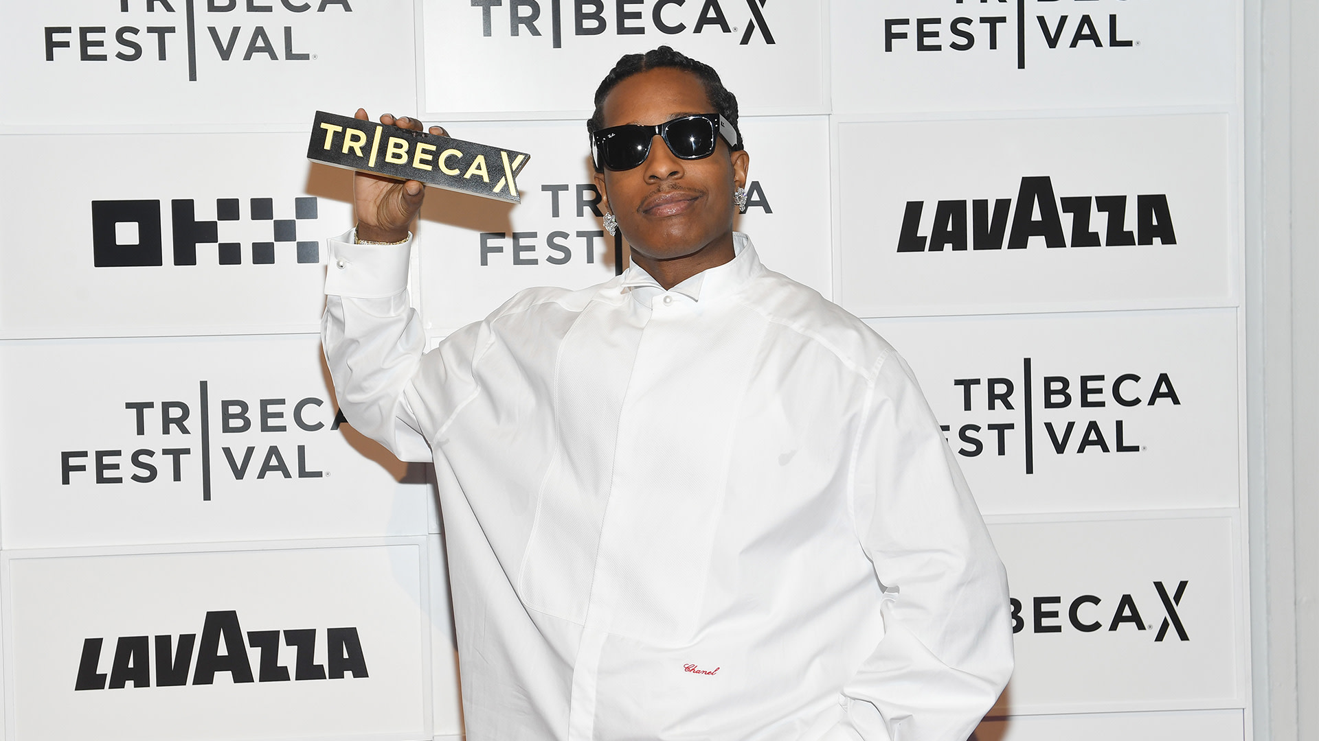 A$AP Rocky in a white shirt and sunglasses at the Tribeca Festival, holding a Tribeca sign.
