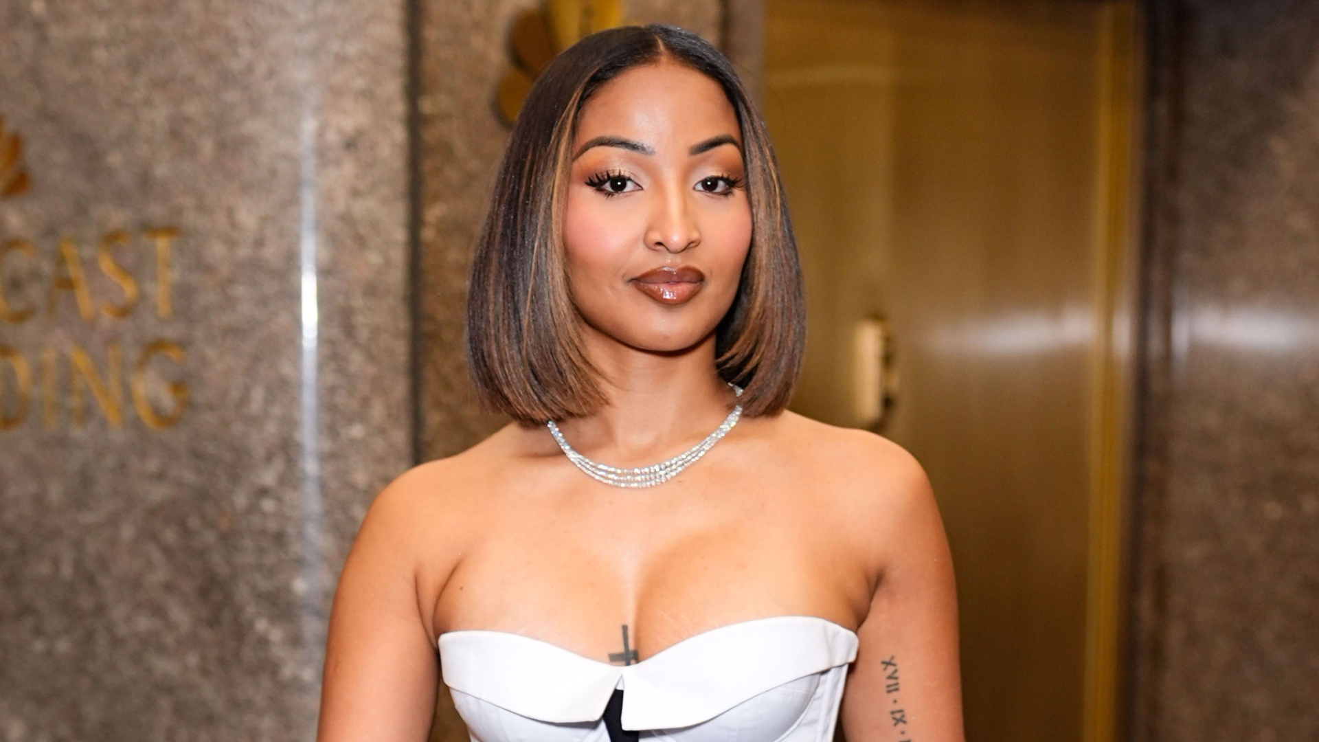 Shenseea with shoulder-length hair in a strapless white dress stands confidently in front of a stone wall.