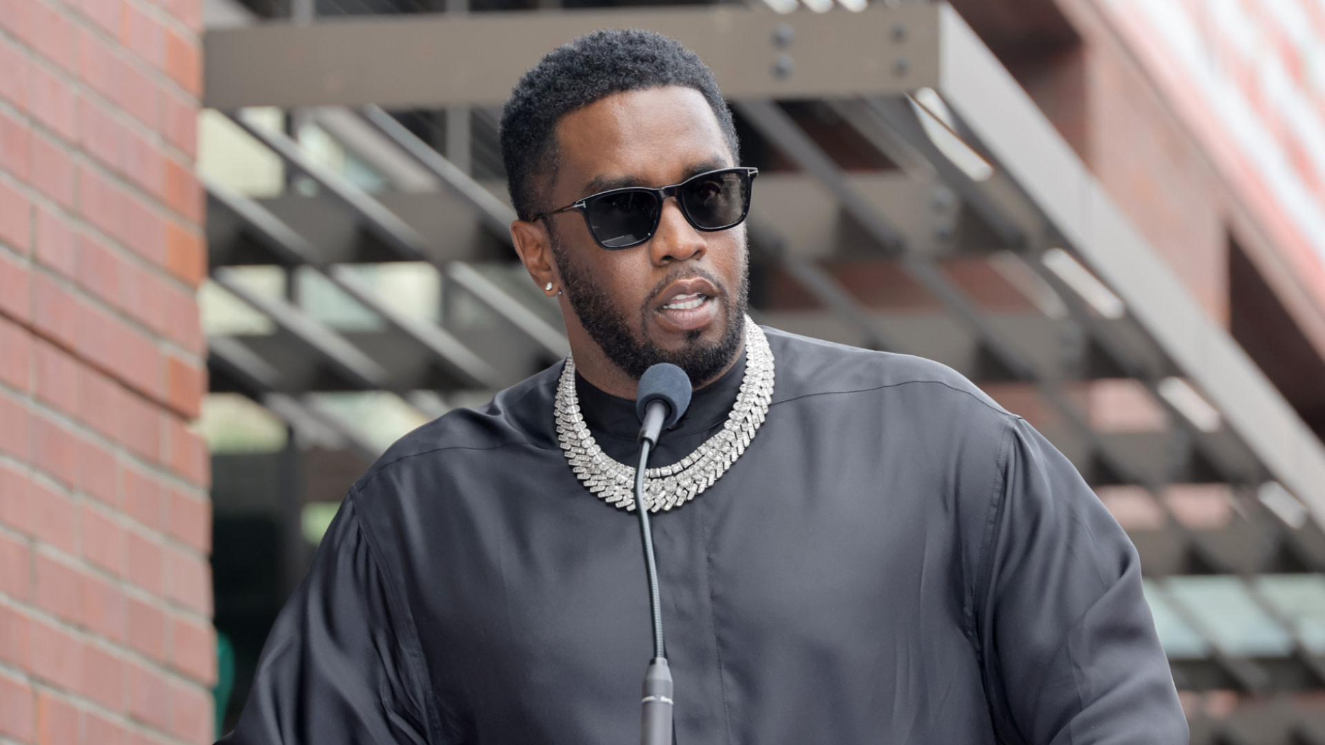 Diddy wearing sunglasses and a black outfit speaks at a podium, with a layered necklace and a brick wall in the background.
