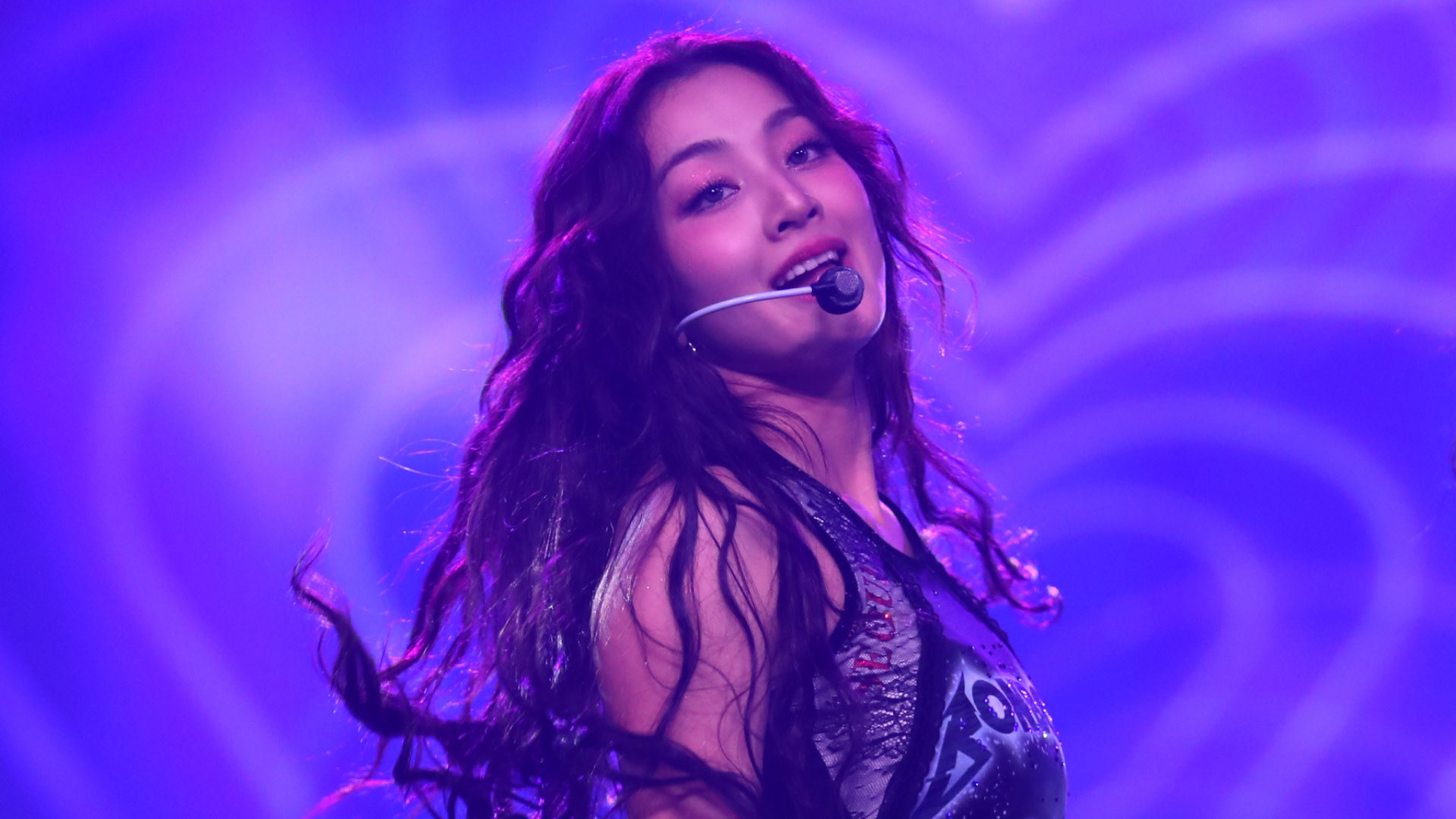 Jihyo with long, wavy hair and a headset microphone sings on stage with a purple heart-themed background.