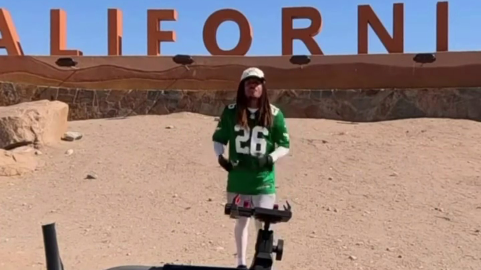 Streamer HMBL Zay in a green football jersey stands in a desert area with a sign reading "CALIFORNIA" in the background.