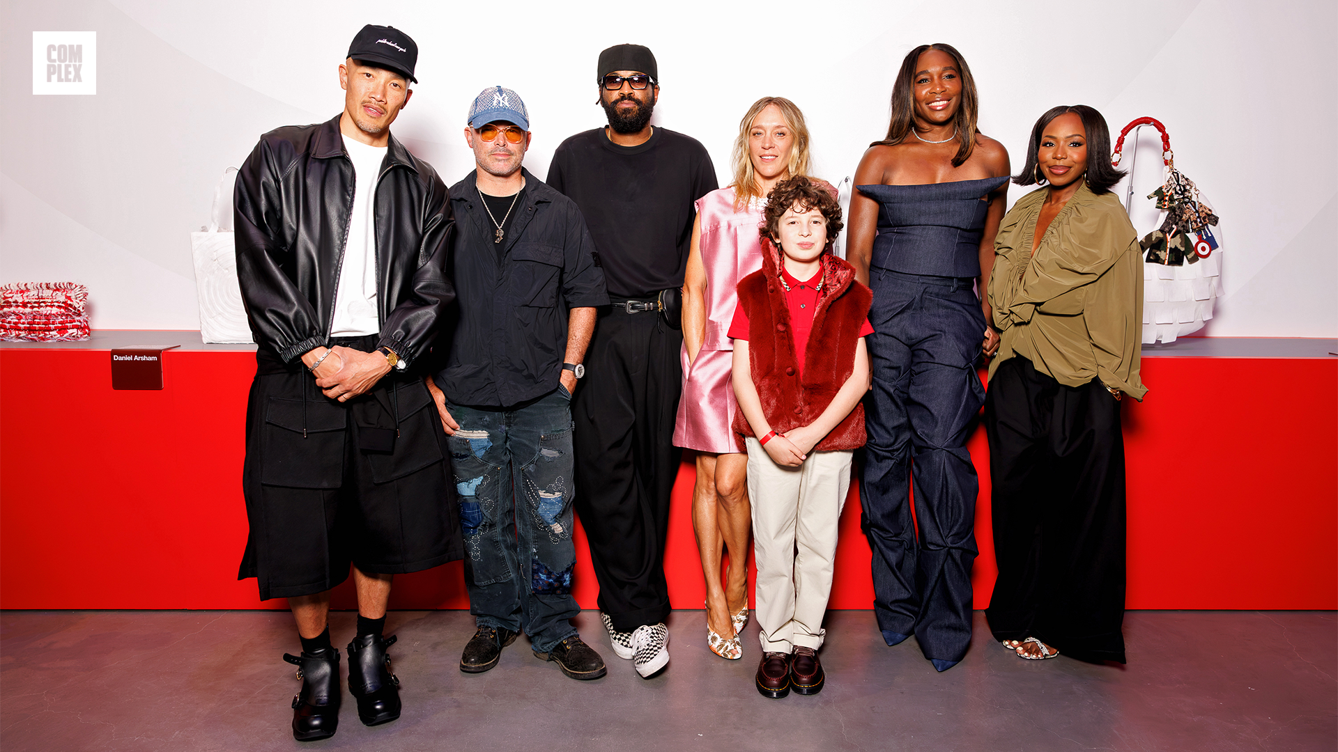 A group of people, including a child in a red outfit, stand in front of a red and white backdrop. They are dressed in stylish, modern clothing.