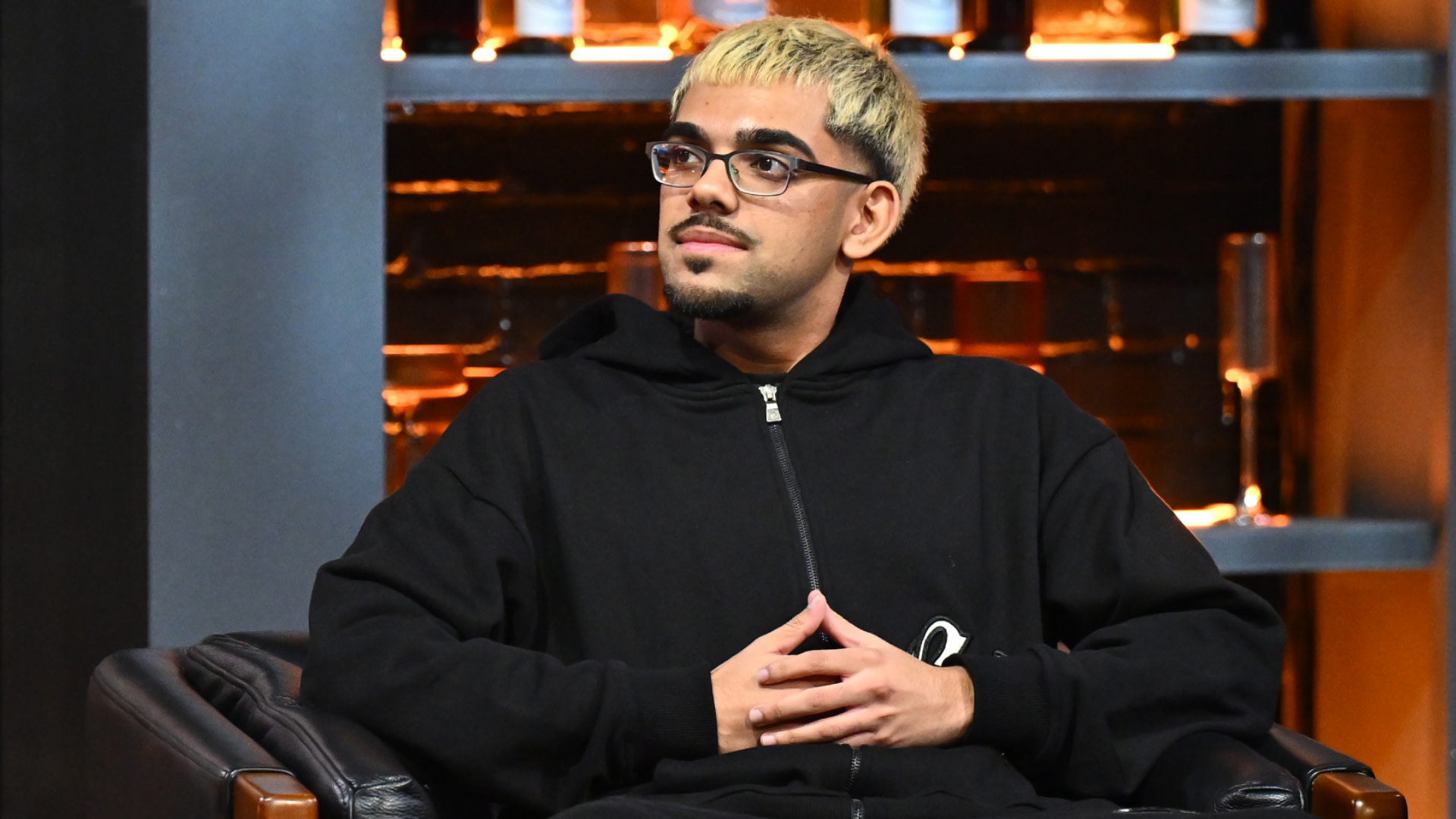N3on with glasses and blonde hair sits in a chair, wearing a black hoodie, against a dimly lit background.