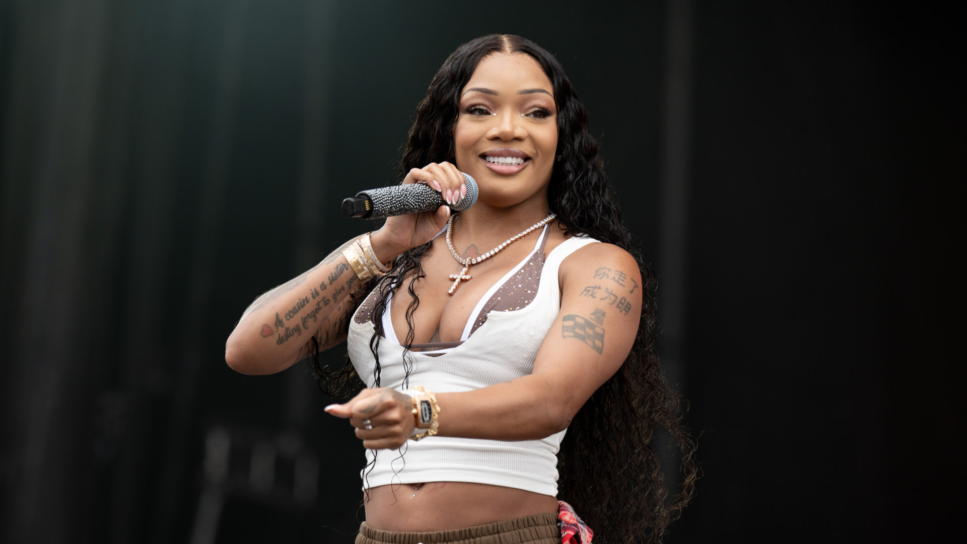 GloRilla with long black hair and tattoos performs on stage, holding a microphone and wearing a white tank top.