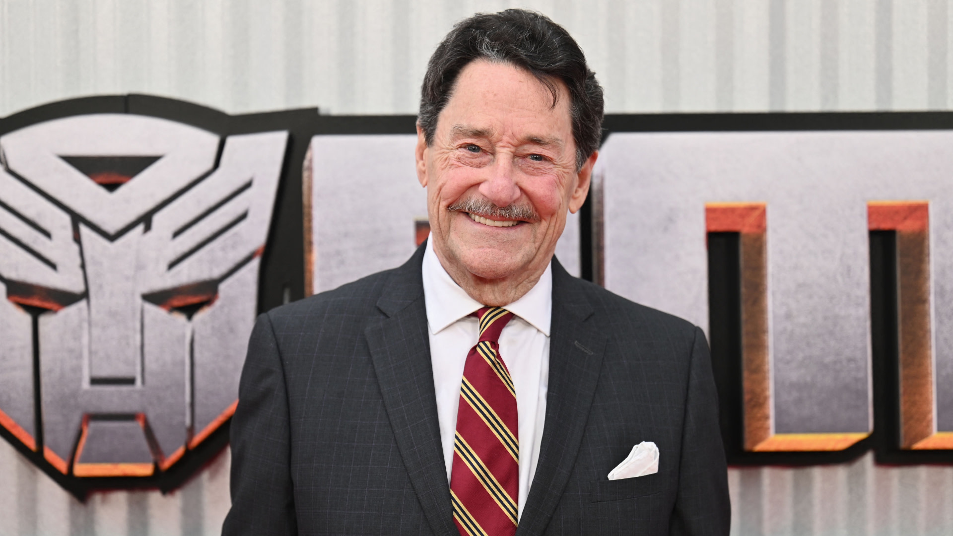 Peter Cullen in a suit and striped tie stands smiling in front of a "Transformers" logo at an event.