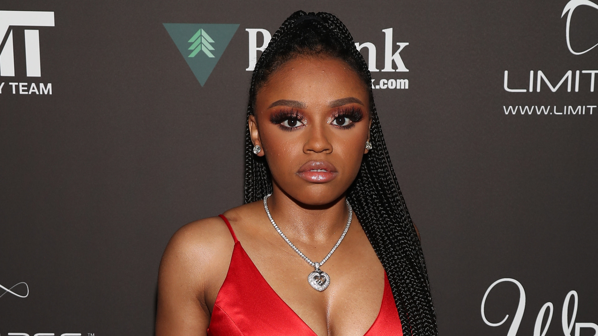 Yaya Mayweather in a red dress with braided hair poses on a red carpet. She wears a heart-shaped necklace and earrings.