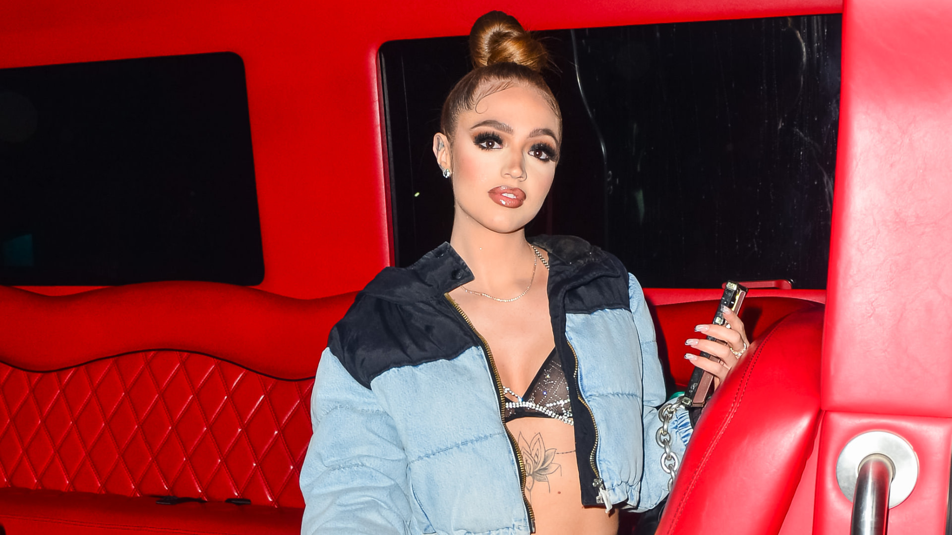Woah Vicky with a top bun hairstyle sits in a red vehicle interior, wearing a denim jacket and holding a phone.