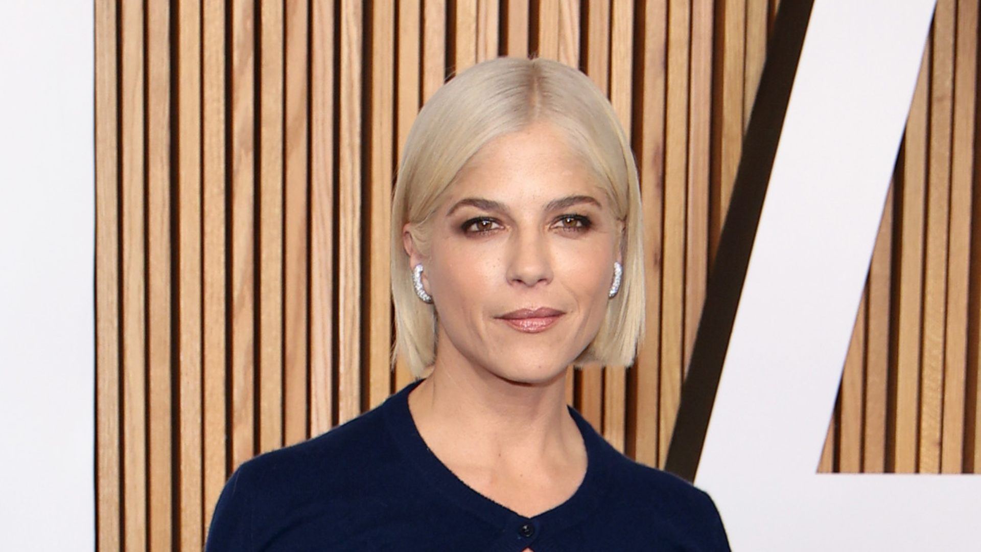 Selma Blair with short blonde hair, wearing a navy top, stands in front of a wooden backdrop.