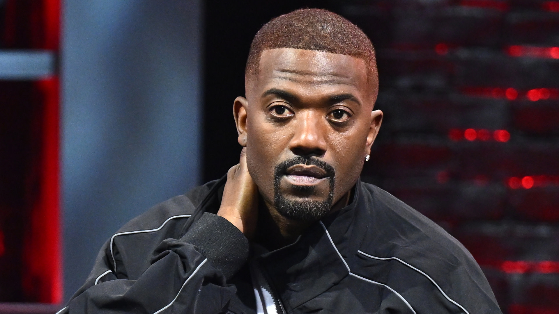 Ray J with short hair and a goatee, wearing a black jacket, sitting with a thoughtful expression.