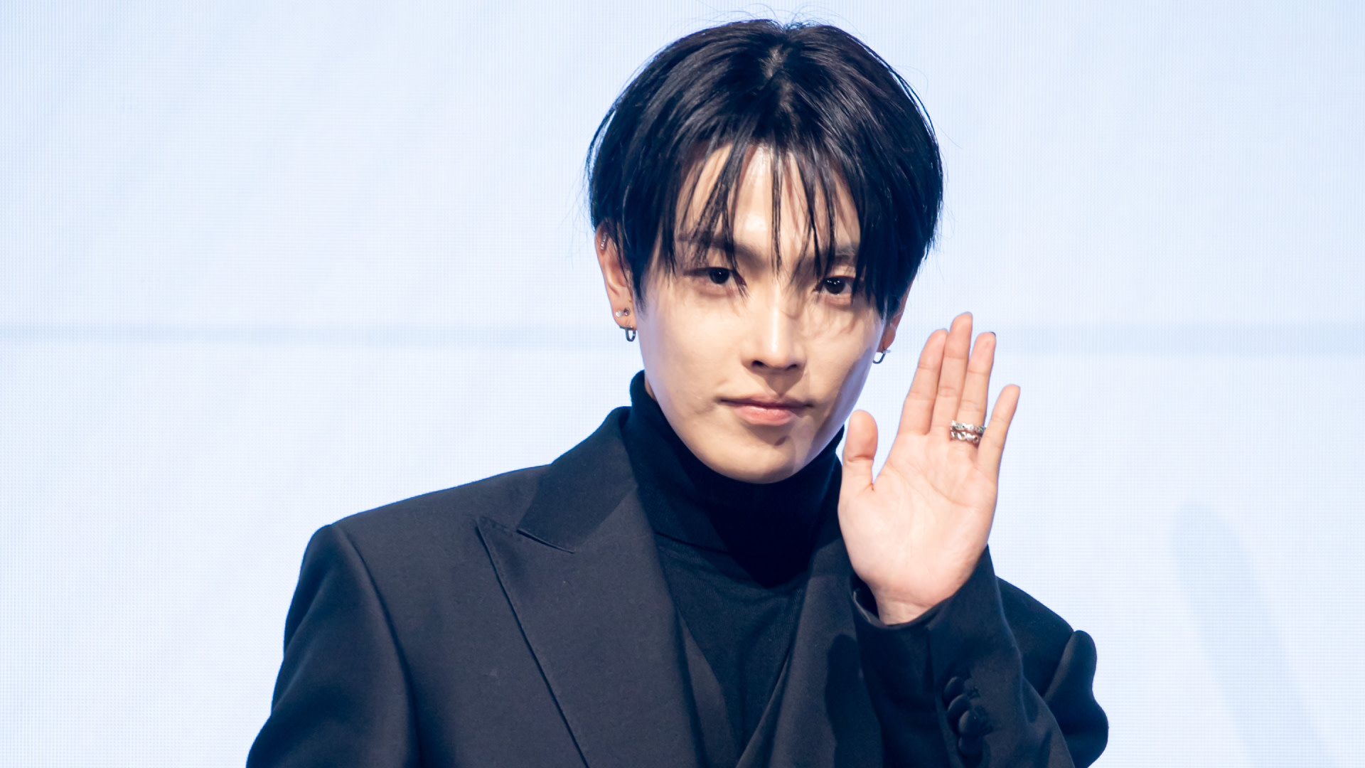 ATEEZ's Hongjoong with dark hair wearing a black suit and turtleneck, raising their hand in a waving gesture.
