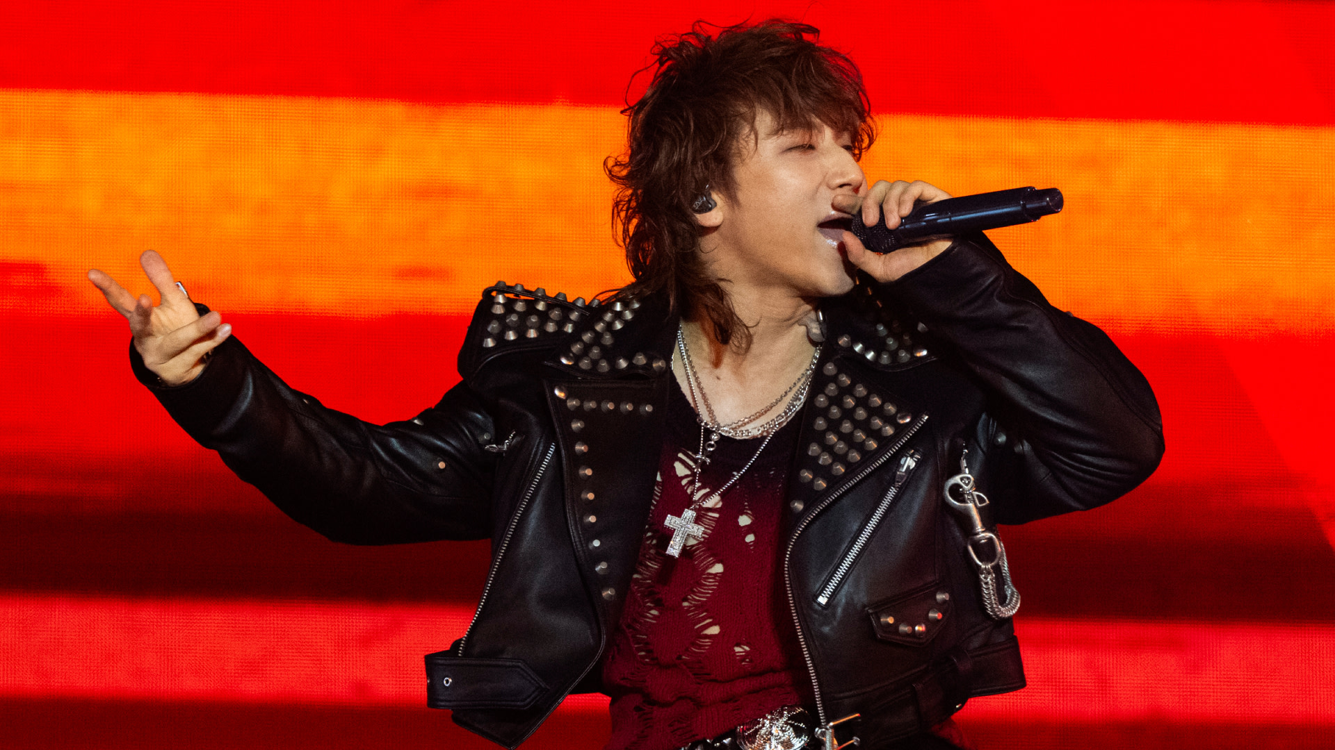 BIGBANG's Daesung in a studded leather jacket sings passionately on stage with a red background.