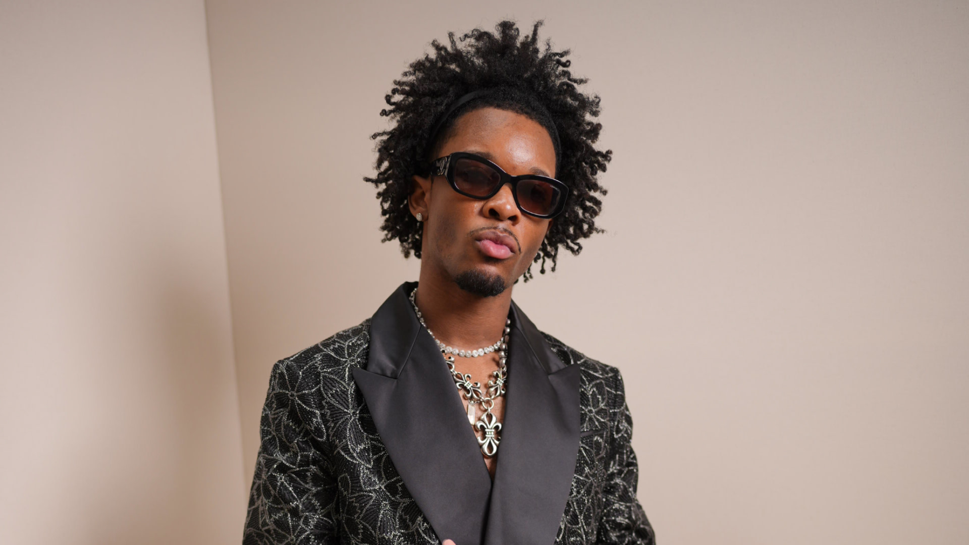 Singer D4vd with curly hair wearing sunglasses, a patterned blazer, and layered necklaces poses confidently against a plain background.
