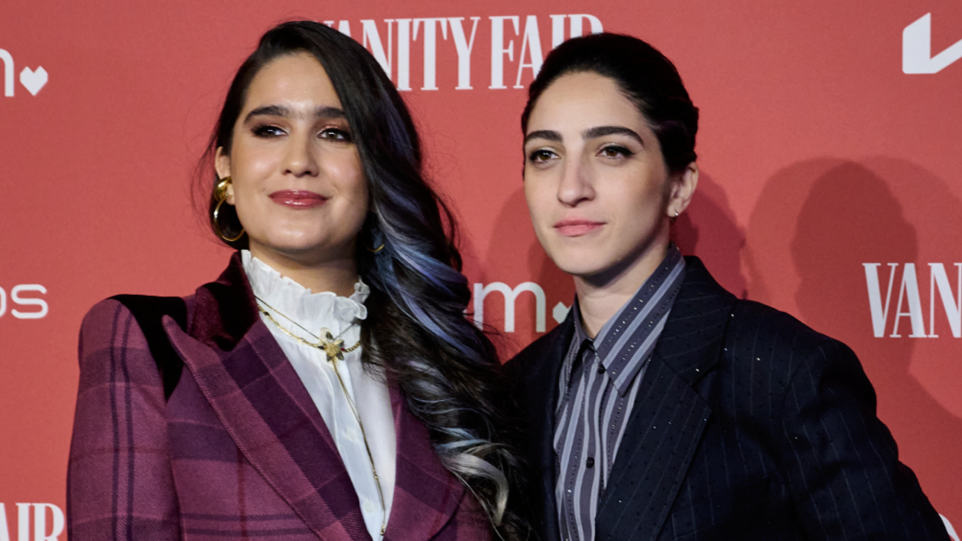 Gemeny Hernández and Emily Estefan posing at an event, dressed in stylish suits against a red backdrop.