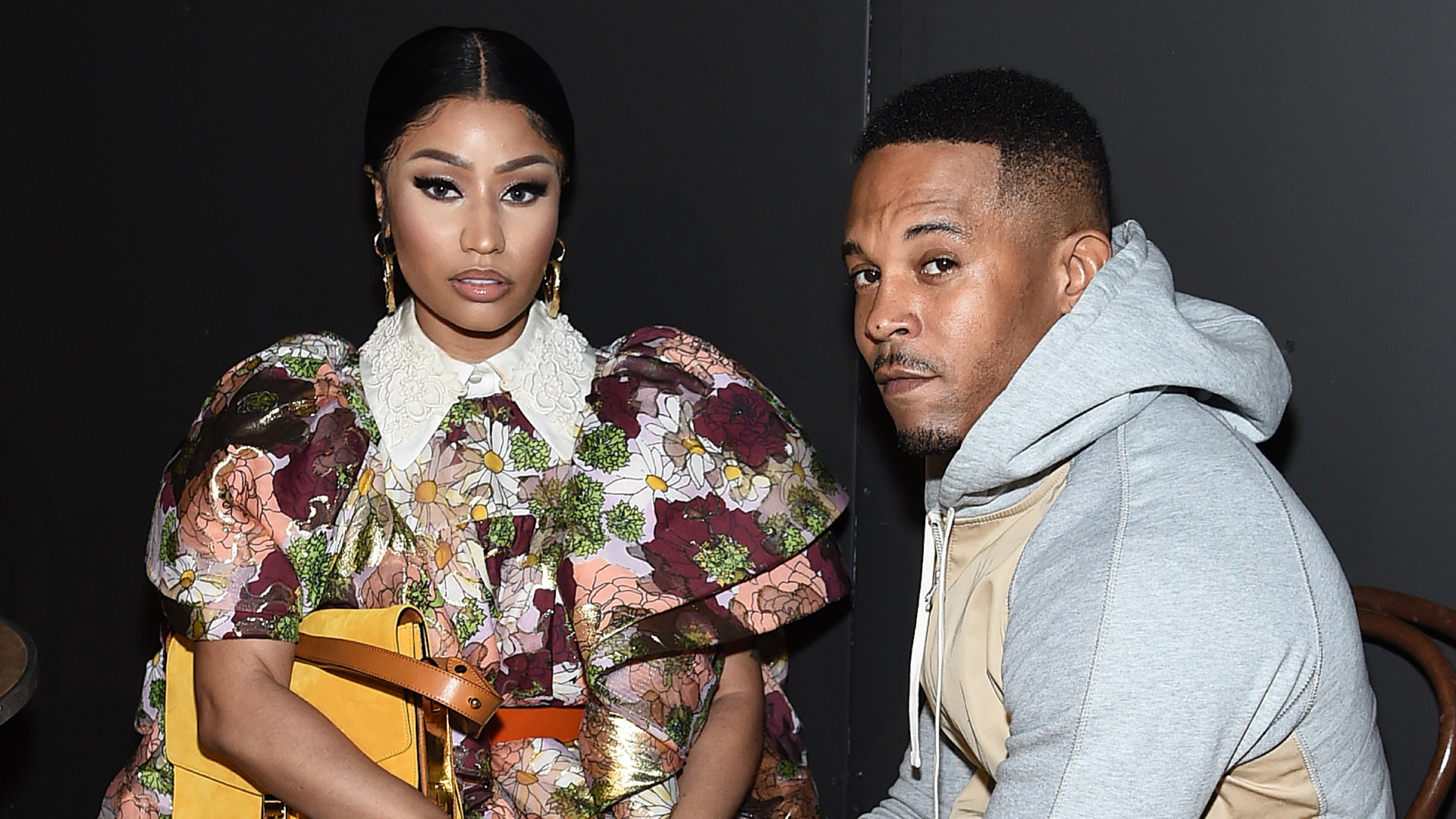 Nicki Minaj in a floral dress with a yellow purse, sitting next to a man in a gray hoodie against a dark background.