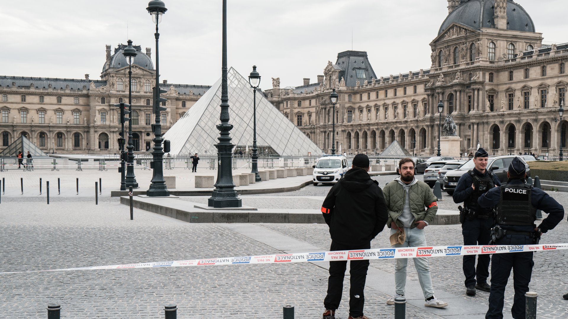 Police officers and two men stand near a cordoned-off area in front of the Louvre Pyramid in Paris. The museum is in the background.