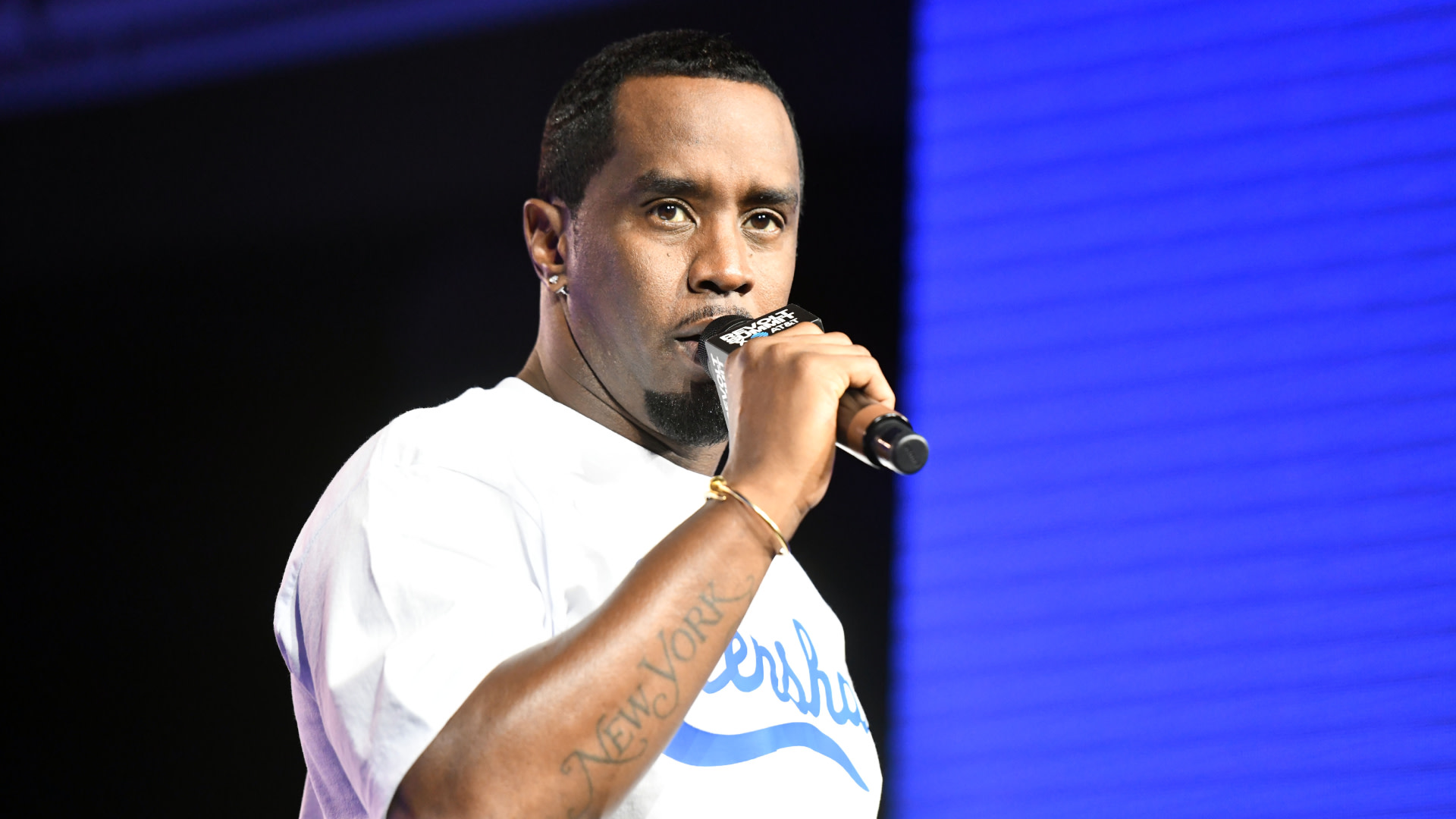 Sean "Diddy" Combs holding a microphone, wearing a white t-shirt, with a tattoo visible on his arm.