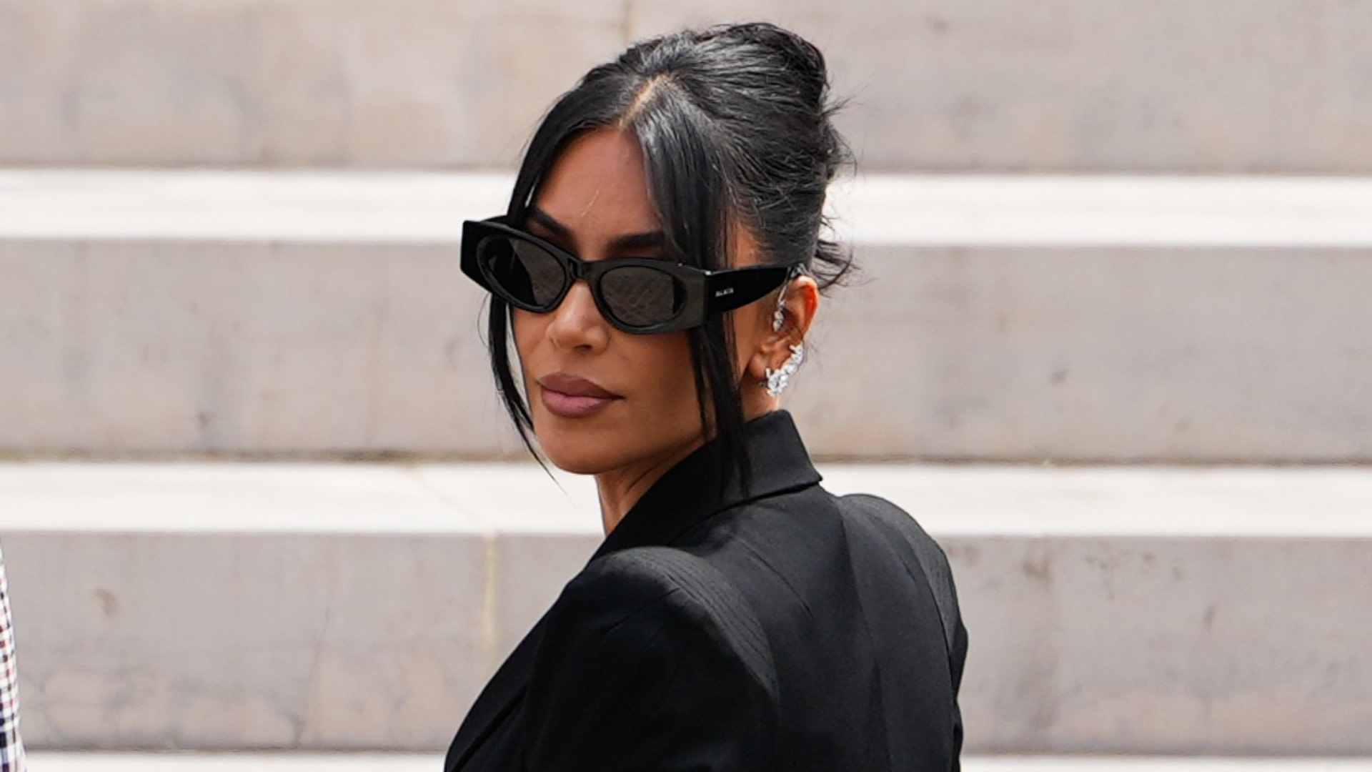 Kim Kardashian wearing black sunglasses and a black outfit, looking over her shoulder with her hair styled up.