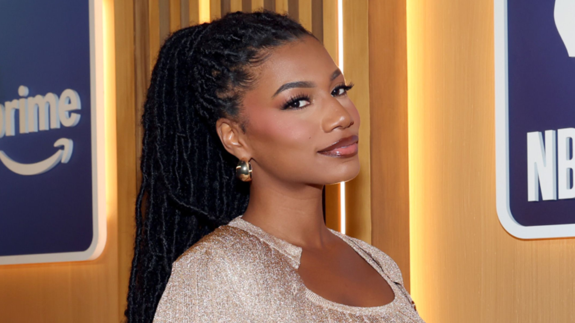Taylor Rooks with long braided hair poses in front of a Prime Video backdrop, wearing a shimmering outfit and earrings.