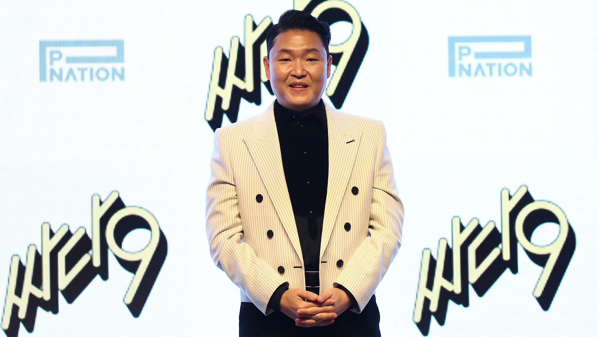 Psy, wearing a striped suit, stands in front of a backdrop with "PNation" and "Happening" logos.