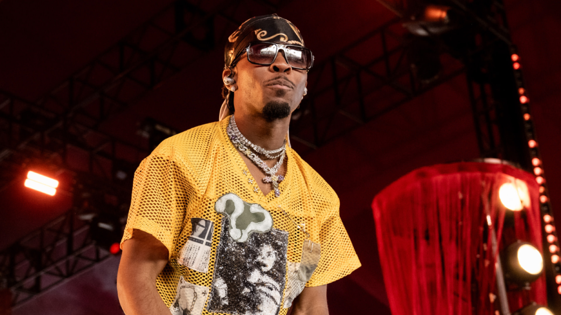 D4vd wearing sunglasses and a yellow mesh shirt on stage, with stage lights in the background.