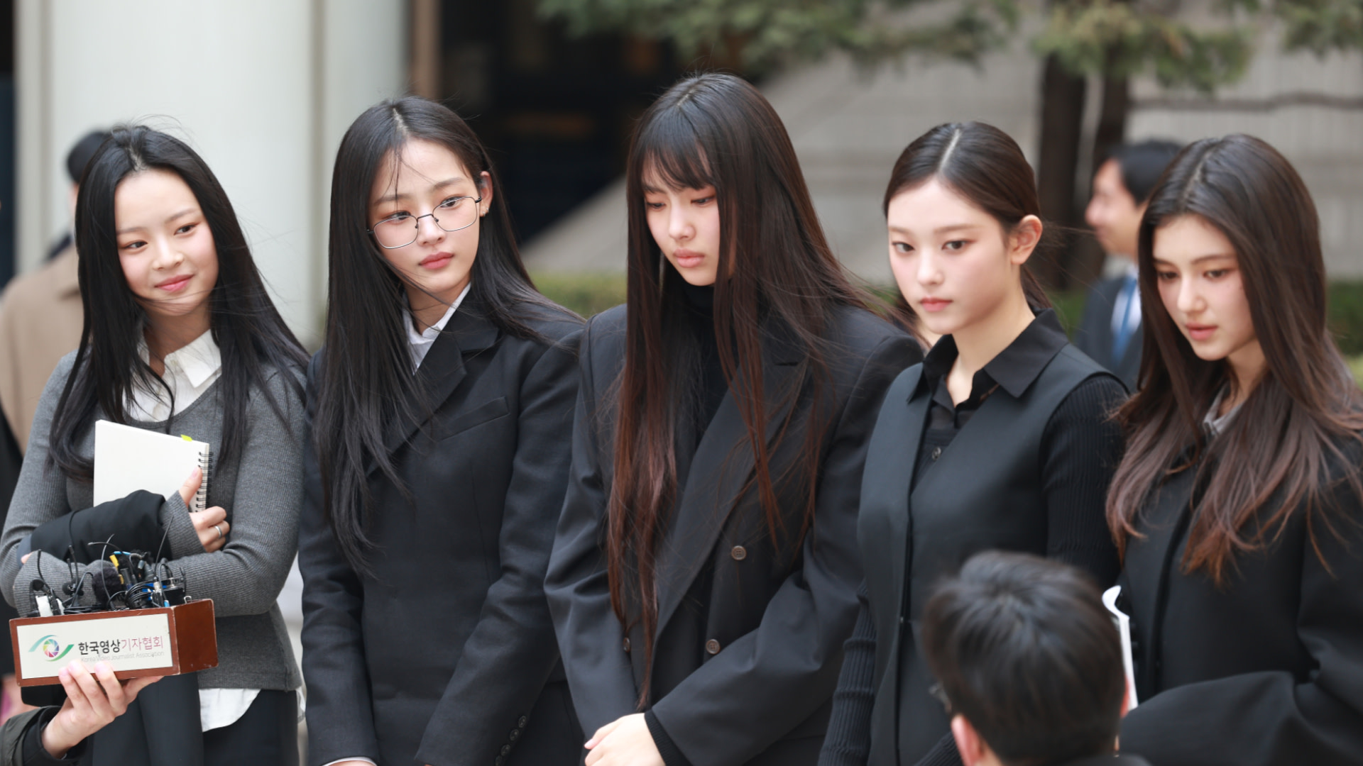 Five young women in black attire stand outdoors, engaged in conversation. One holds a notebook and another wears glasses.